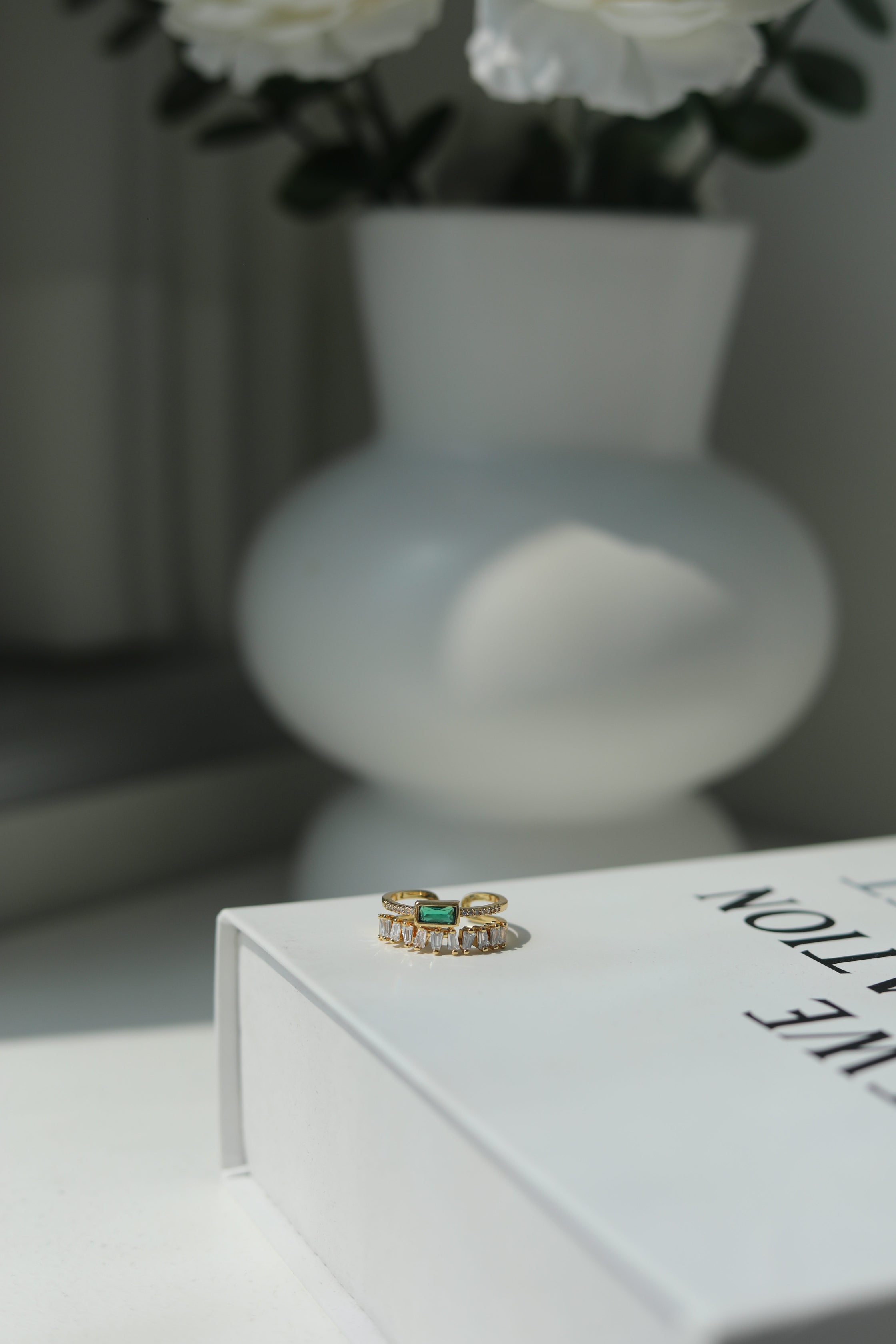 Gold ring with green gemstone on a book with a blurred vase and flowers in the background
