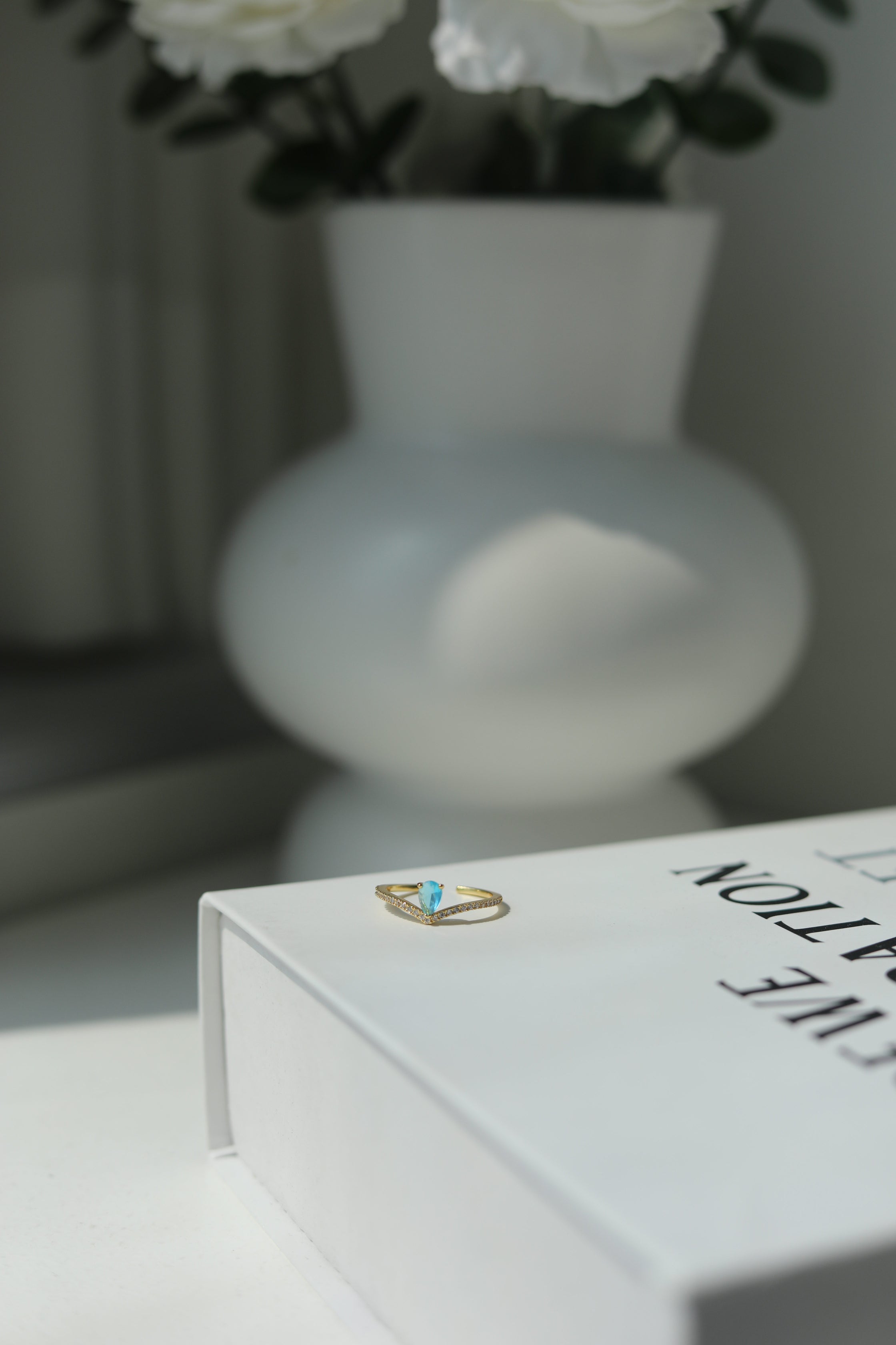 Close-up of a delicate golen ring with a light blue gemstone and a blurred vase and flowers in the background