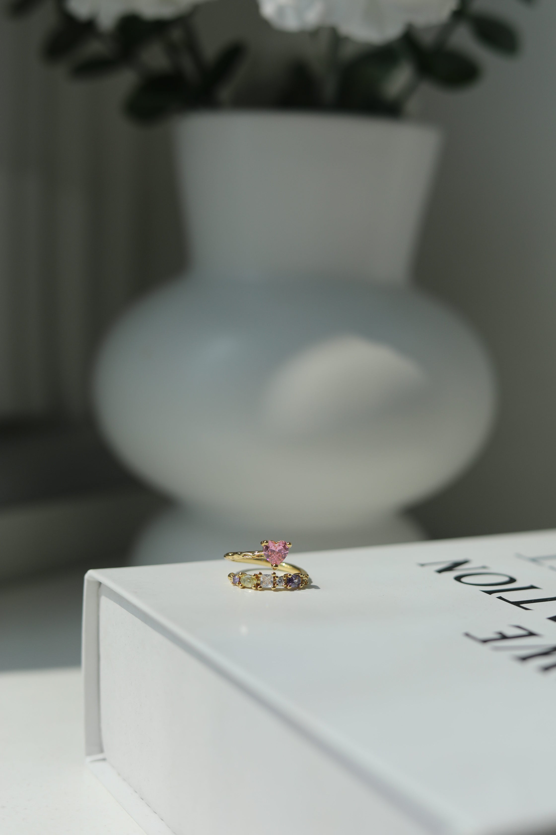 Cute ring with pink and lavender gemstones on a white surface with a blurred vase and flowers in the background