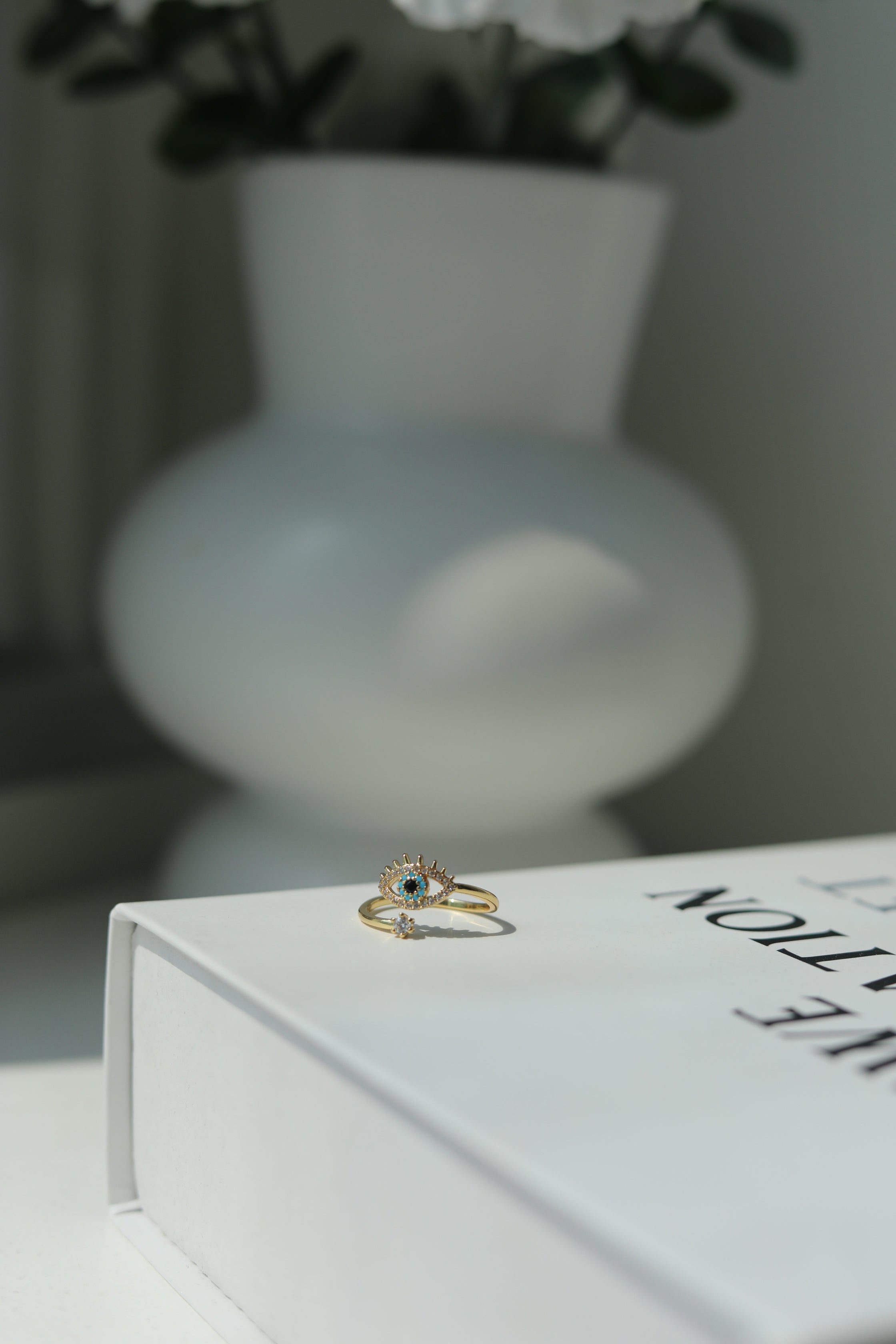 Gold evil eye stainless steel ring with a blue gemstone on a white book with a blurred vase and flowers in the background