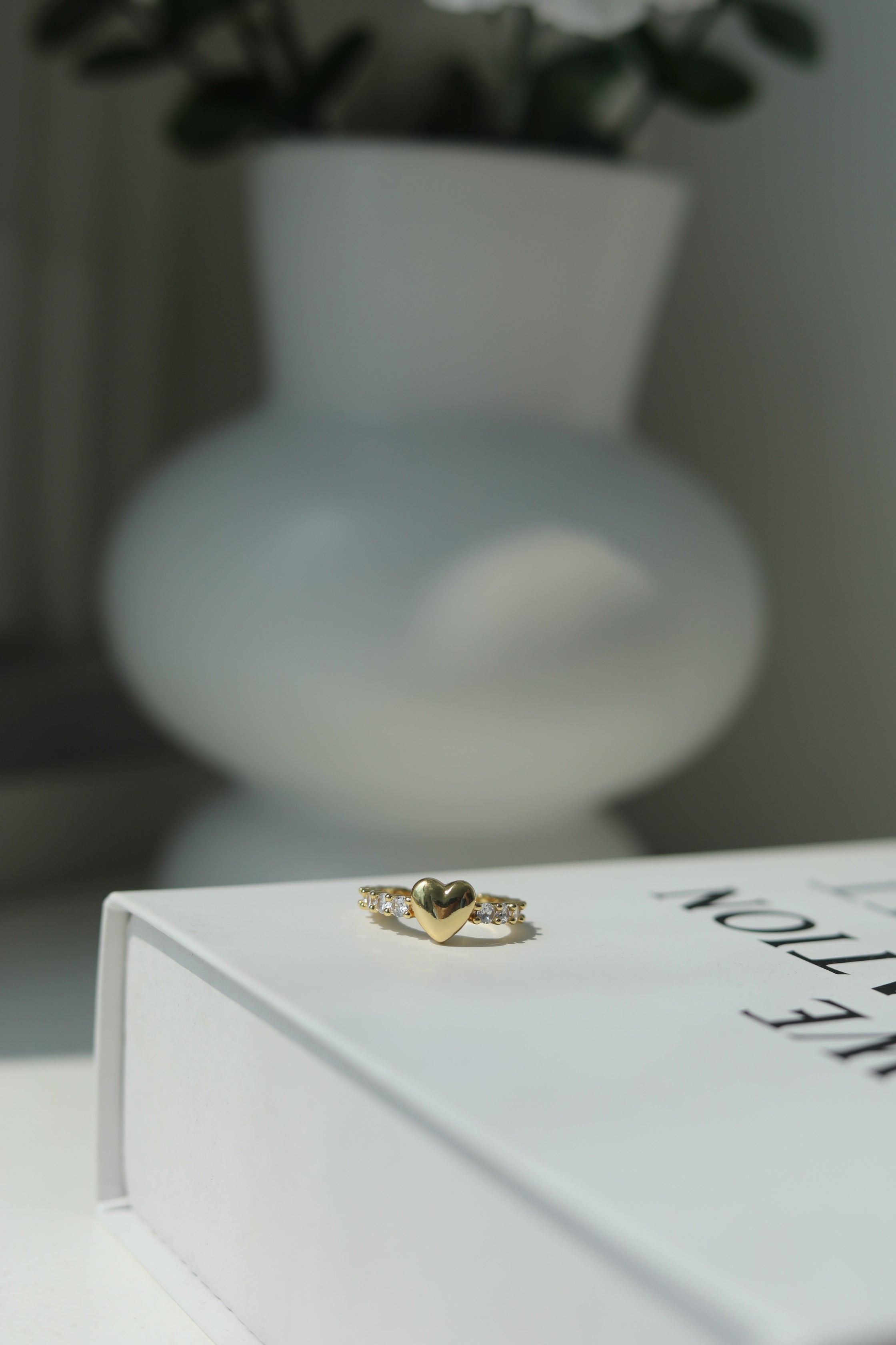 Adjustable gold ring with heart design on a white box with a blurred vase in the background