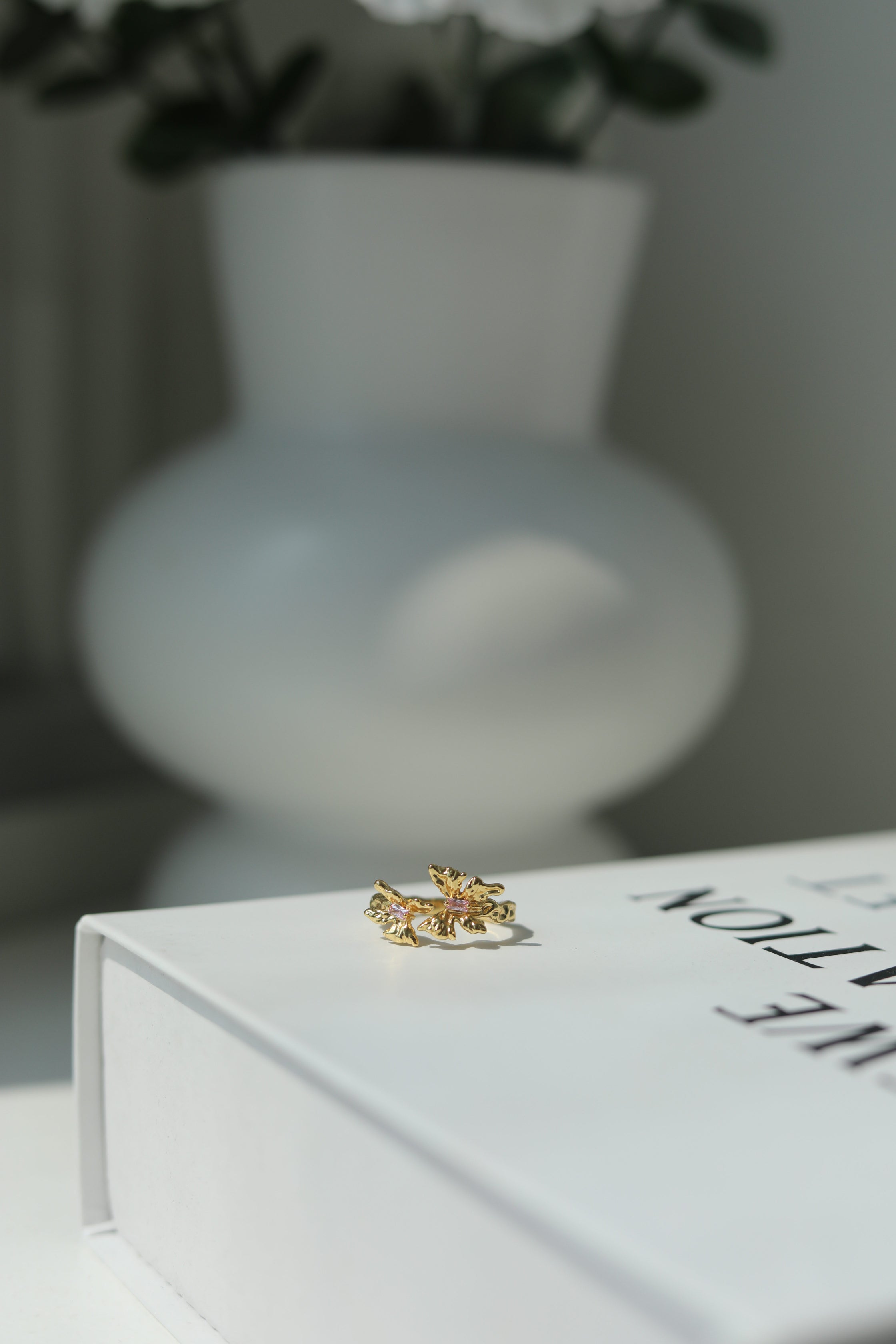 Gold ring with gemstones on a white surface with a blurred vase in the background