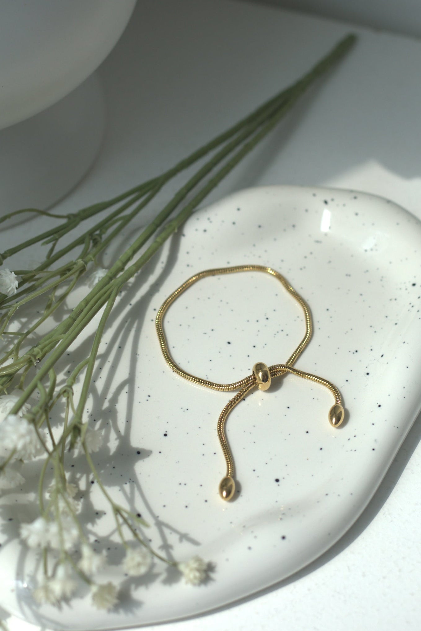 Gold bracelet on a white ceramic dish with small white flowers and green leaves.