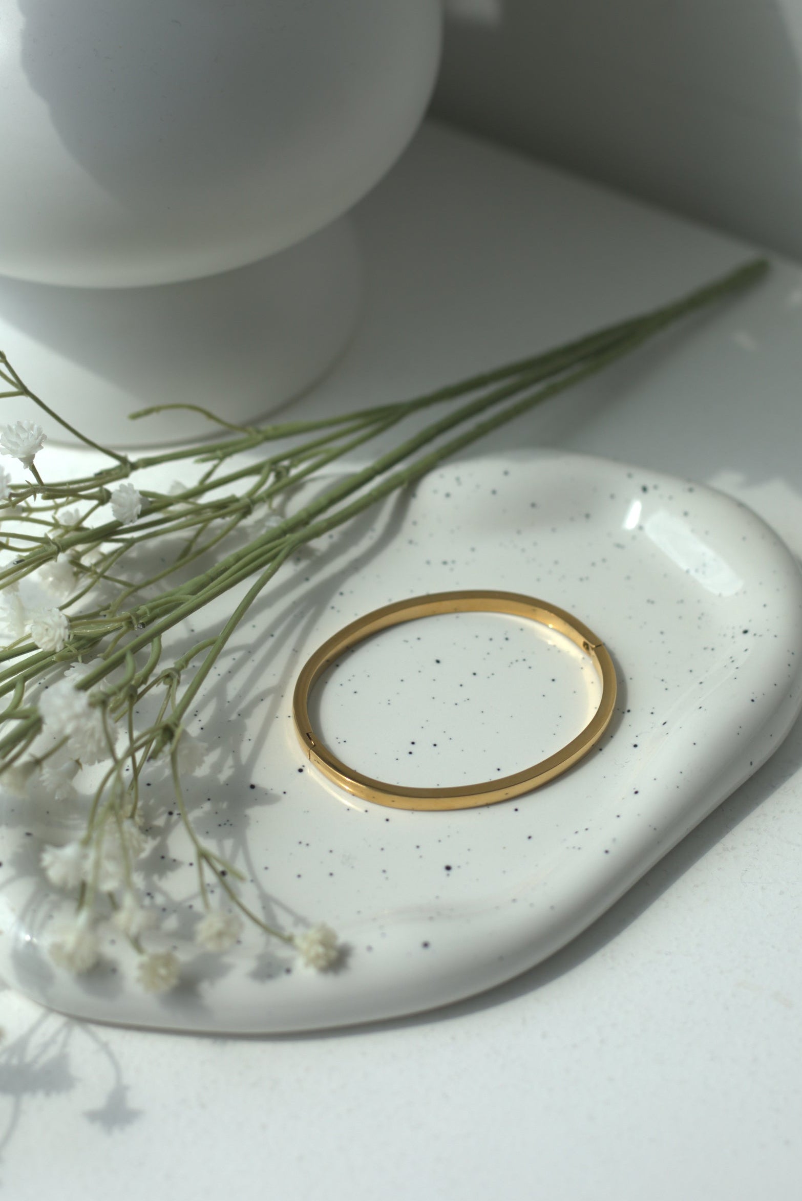 White vase with flowers on a white surface next to a small round white dish with a gold bracelet.