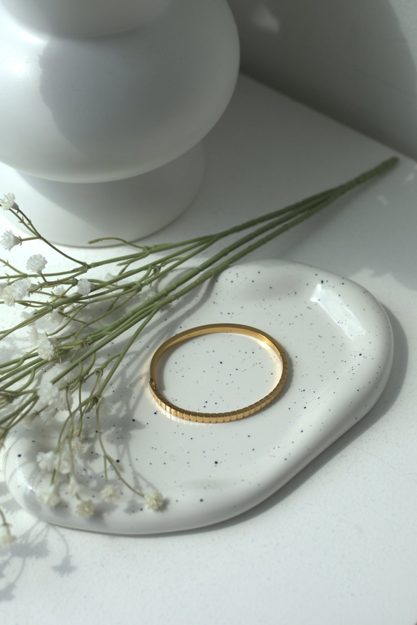 White vase with flowers on a white surface with a gold bracelet and small plant.