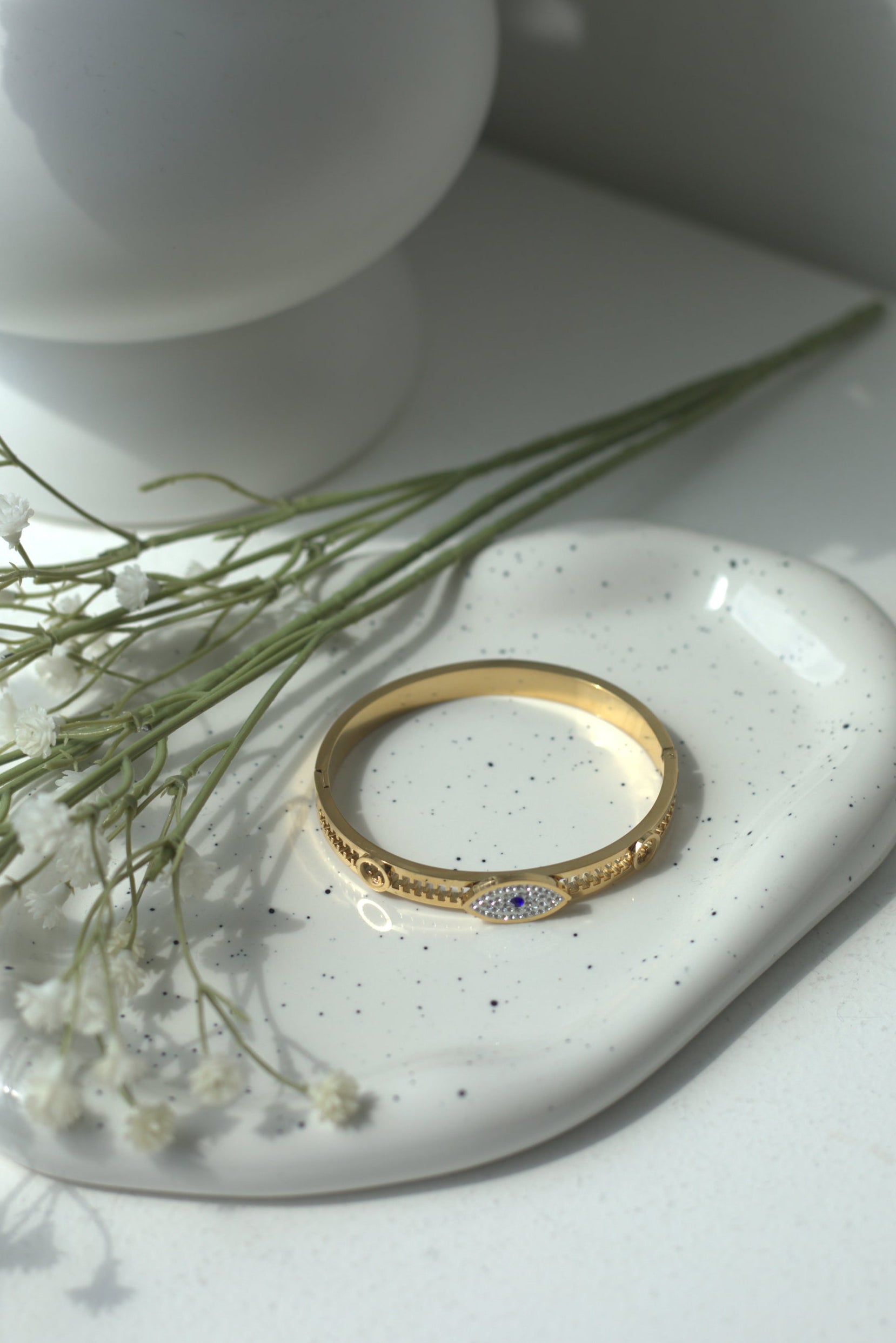 Gold evil eye jewelry bracelet on a white ceramic dish with a white vase and greenery in the background