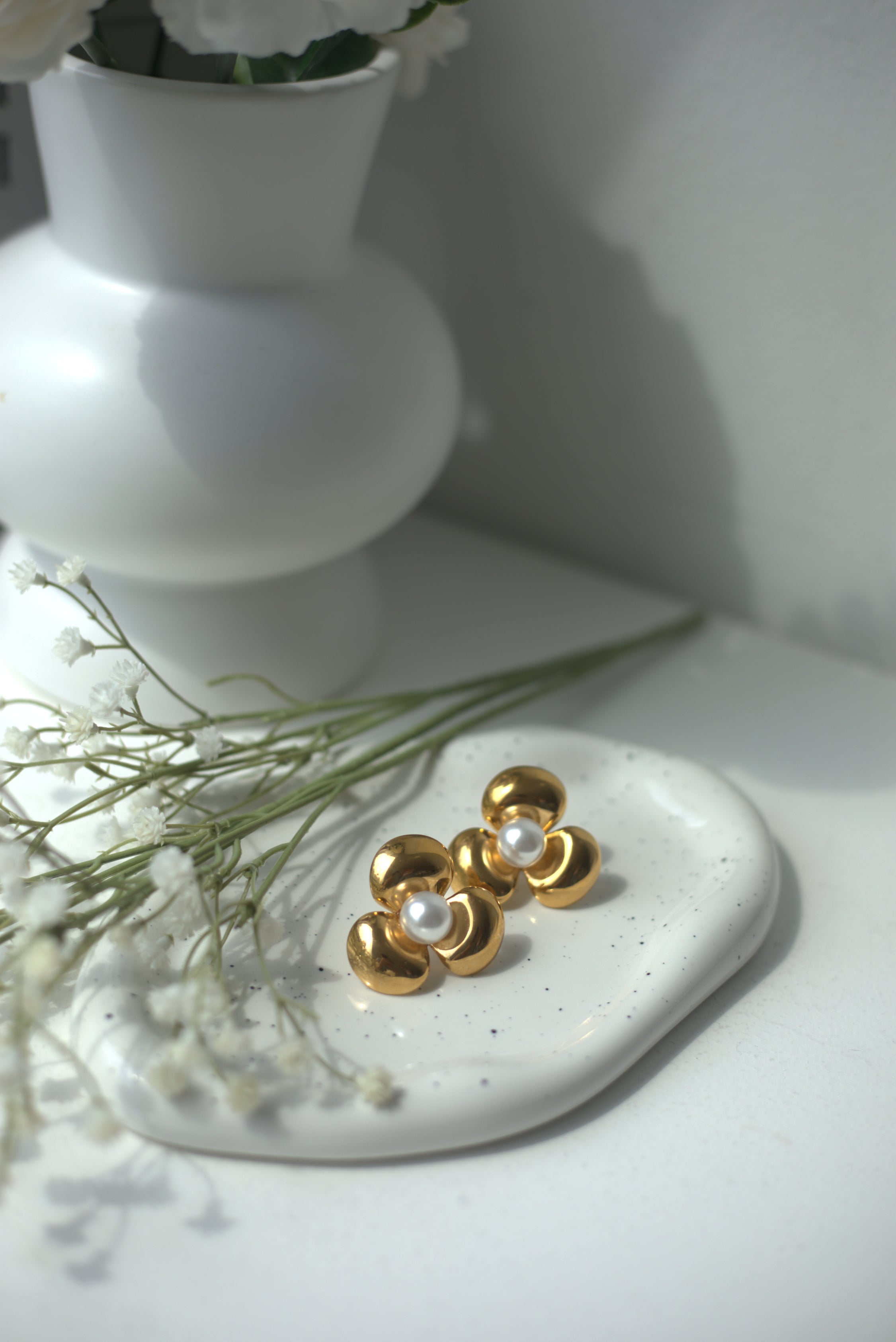 Big gold earrings with pearls on a white surface with a vase and flowers in the background