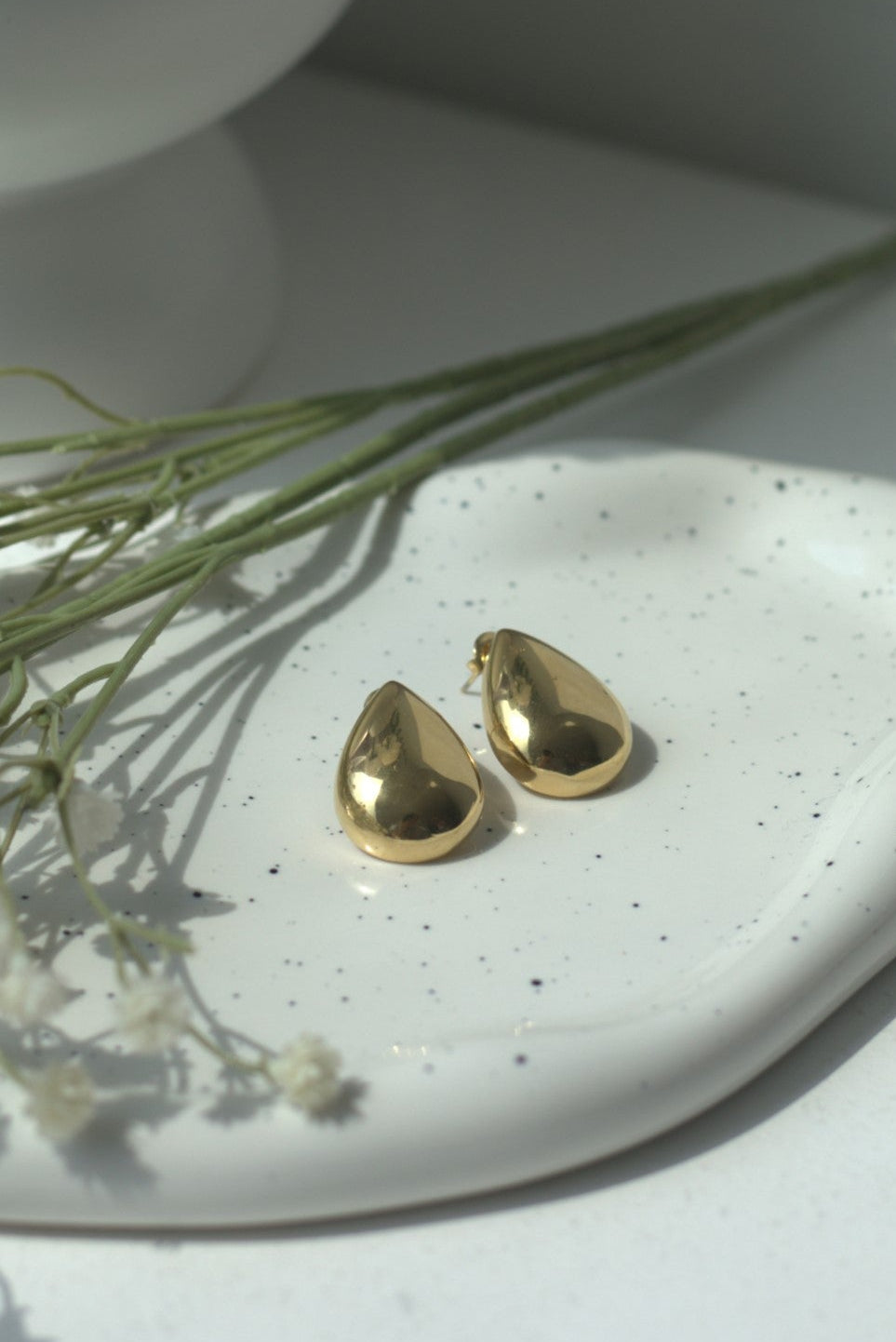 Gold teardrop earrings on a white surface with dried flowers