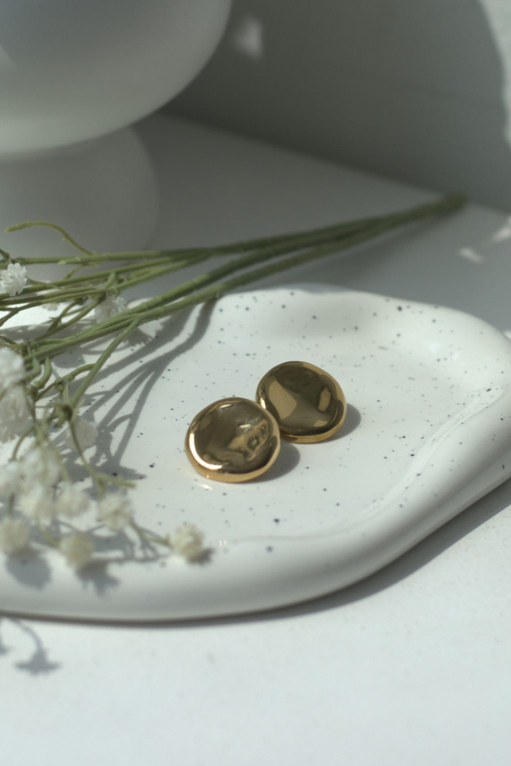 Gold button earrings on a white ceramic dish with small flowers, against a light background