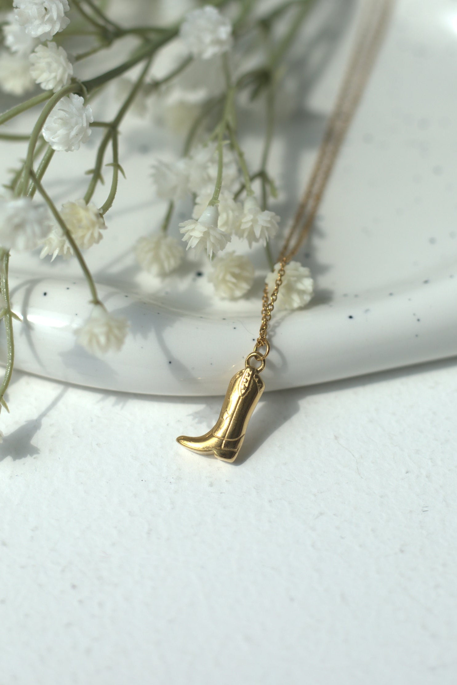 Gold cowboy boot necklace with a small pendant on a white surface with baby's breath flowers.