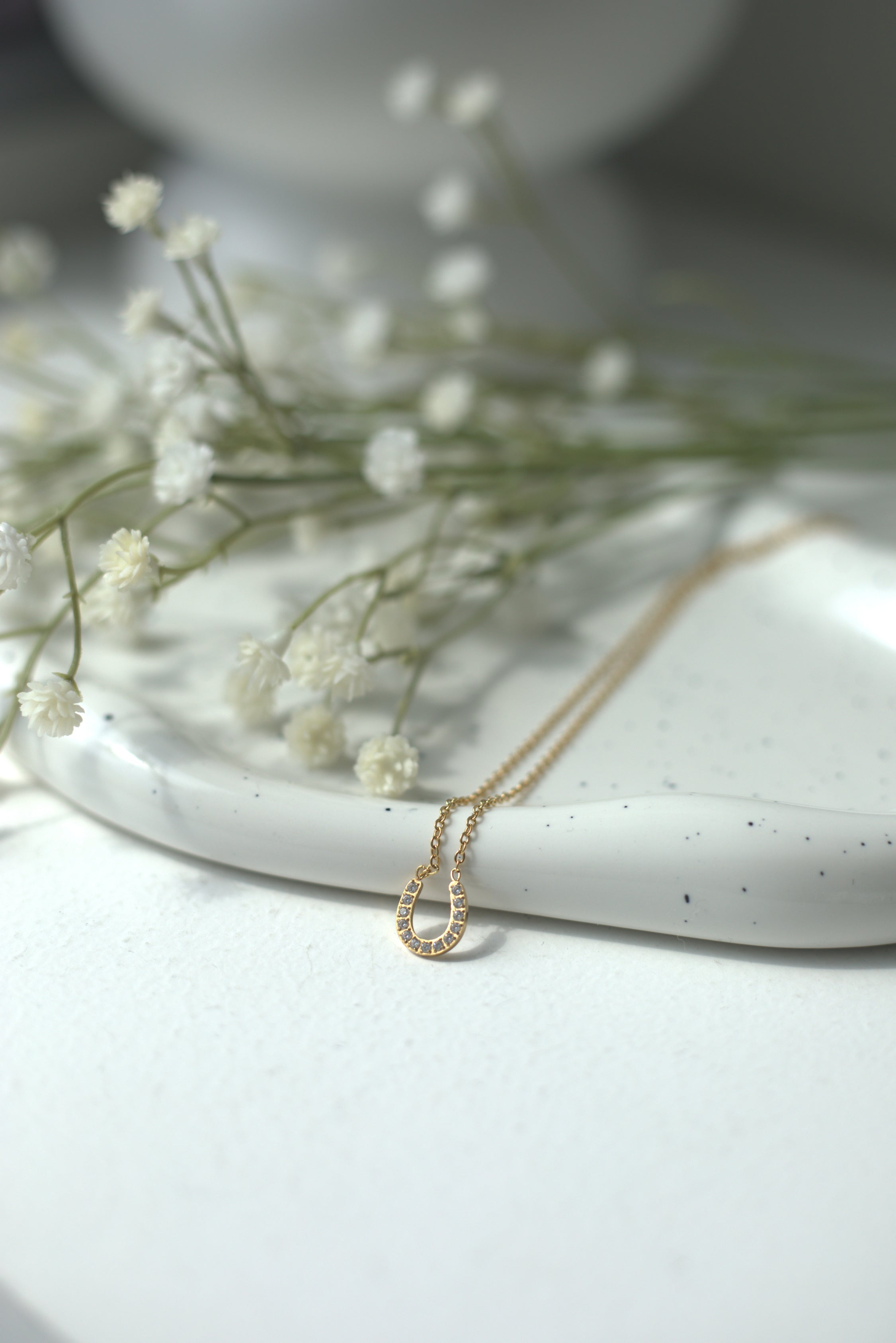 Gold delicate horseshoe necklace on a white surface with small white flowers. For good luck & prosperity