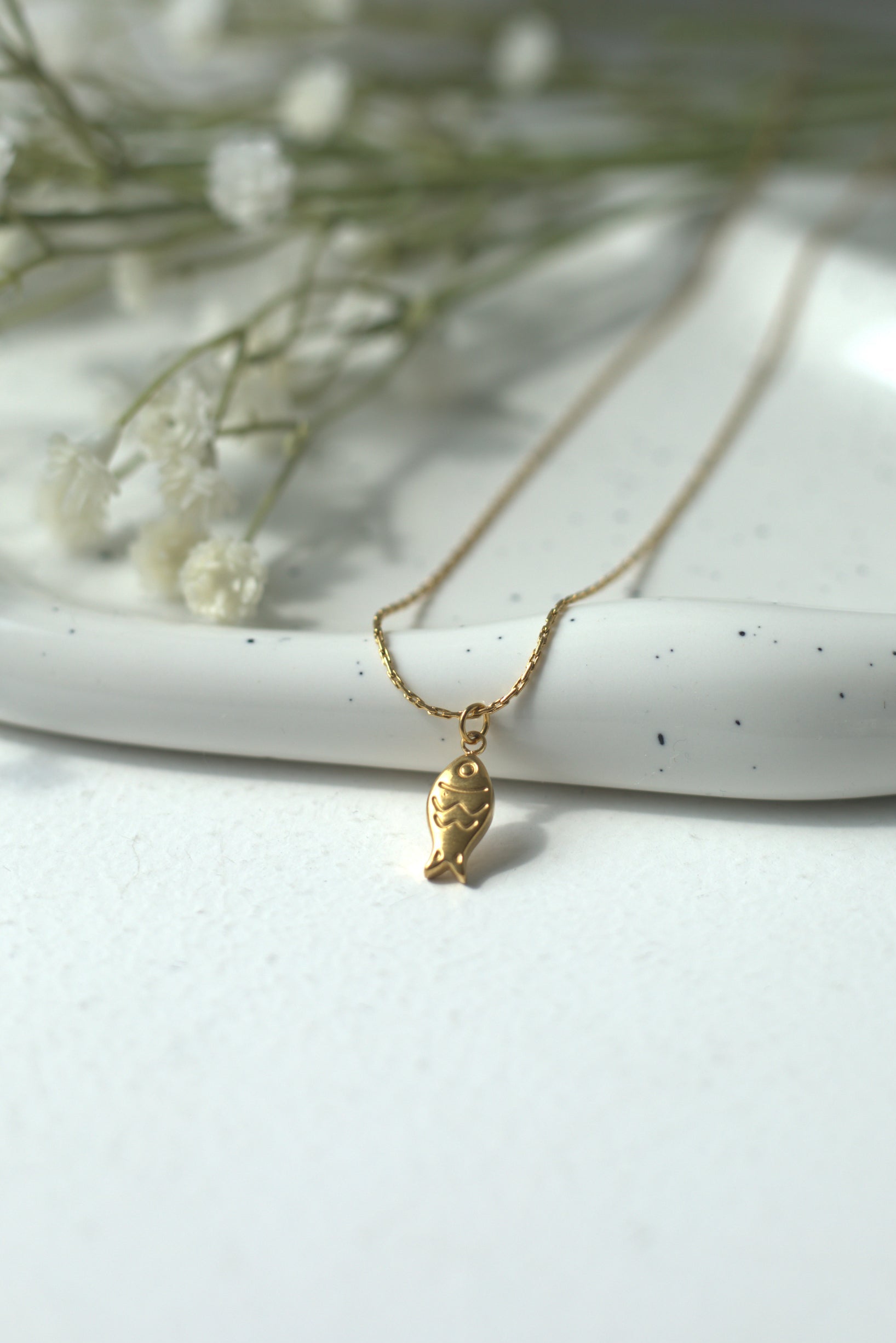 Gold stainless steel necklace with goldfish pendant on a white surface with small white flowers
