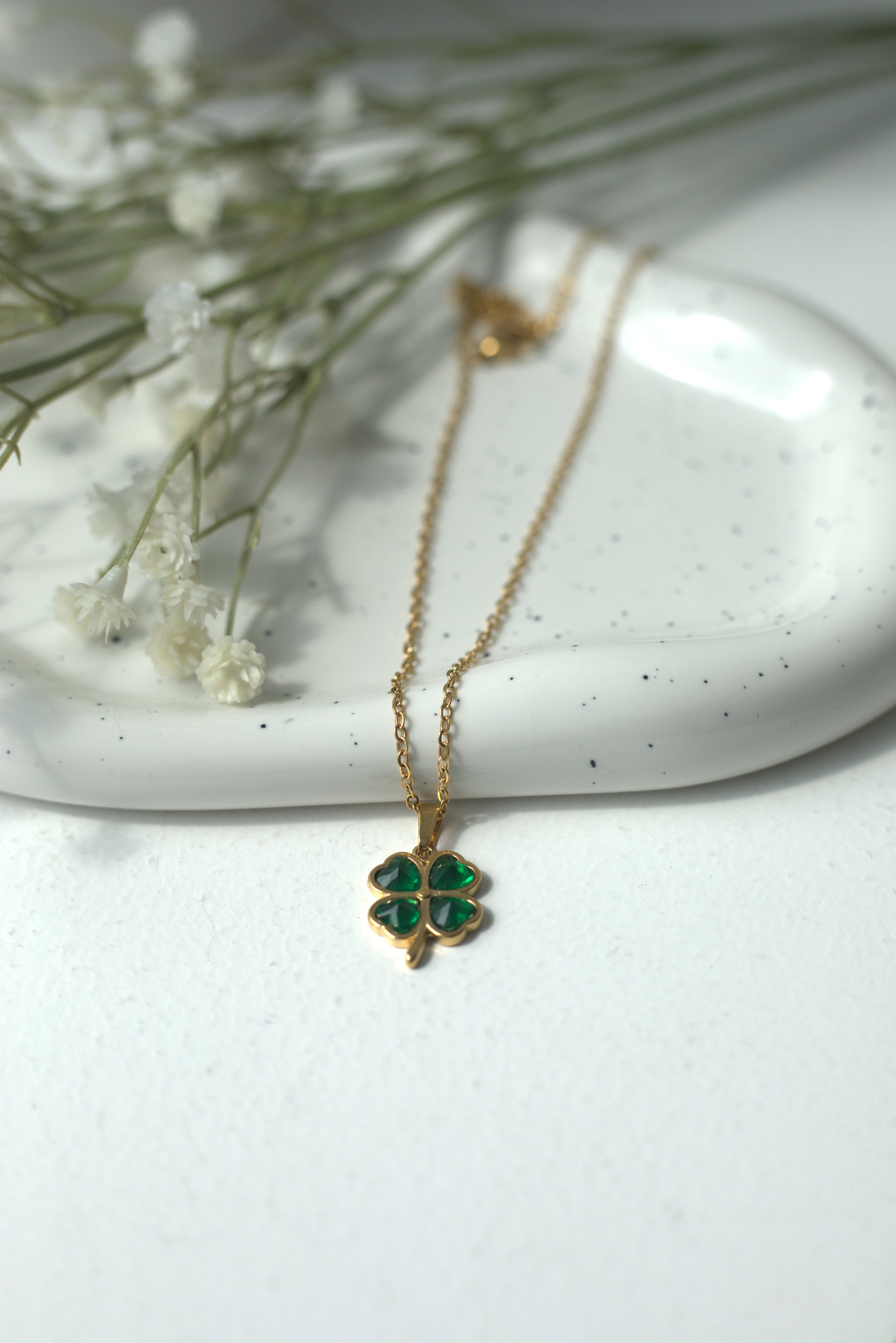 Gold necklace with a green four-leafed-clover pendant on a white surface with dried flowers.