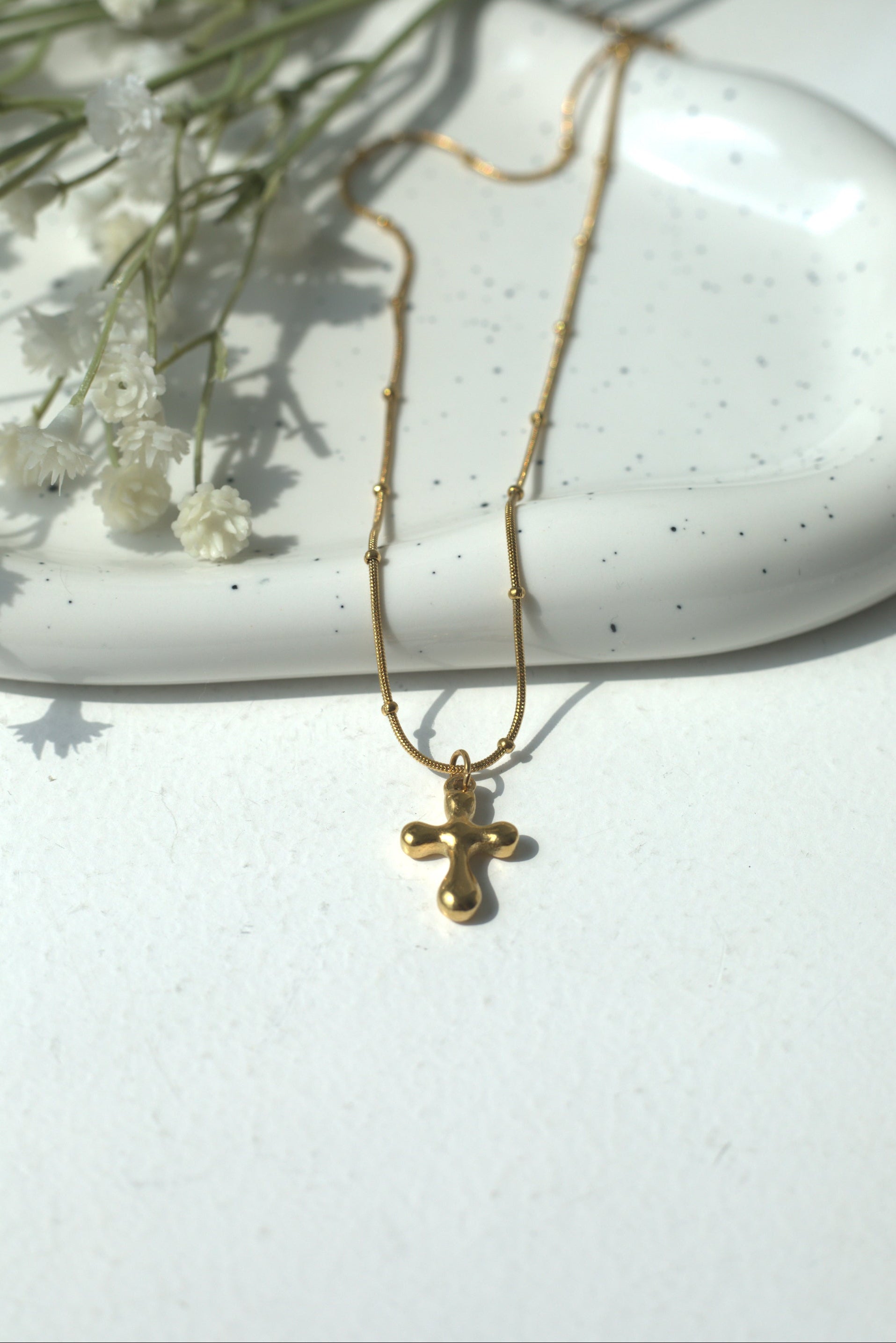 Gold necklace with a stainless steel religious cross pendant on a white surface with dried flowers.