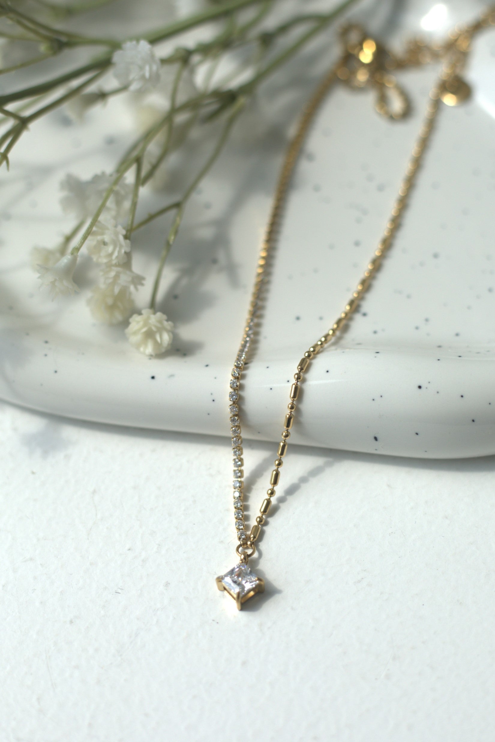Gold necklace with a diamond pendant on a white surface with dried flowers.