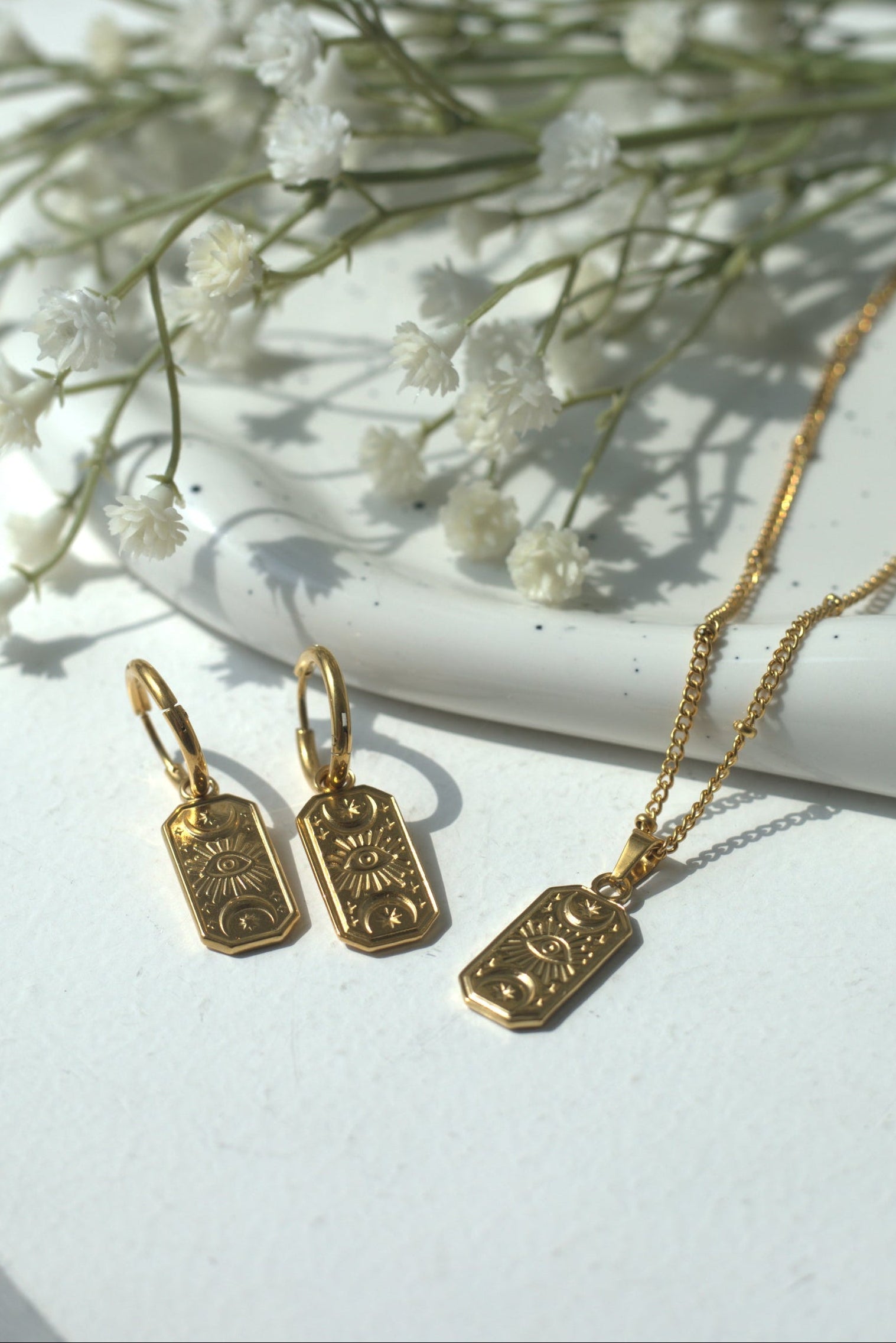 Gold evil eye jewelry set including necklace and earrings on a white surface with small white flowers. Stainless steel