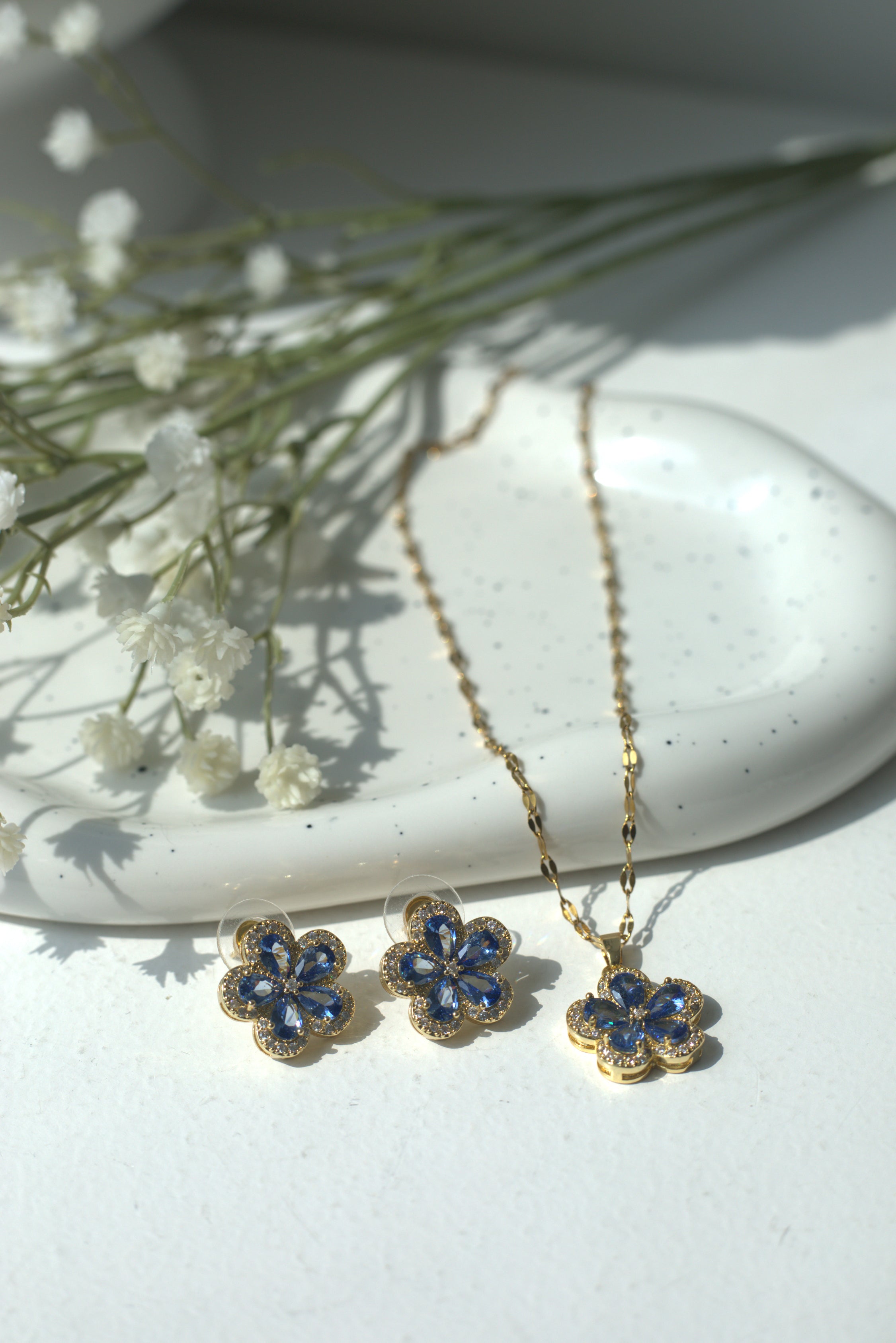 Set of gold necklace and earrings with blue flowers on a white surface with baby's breath flowers.