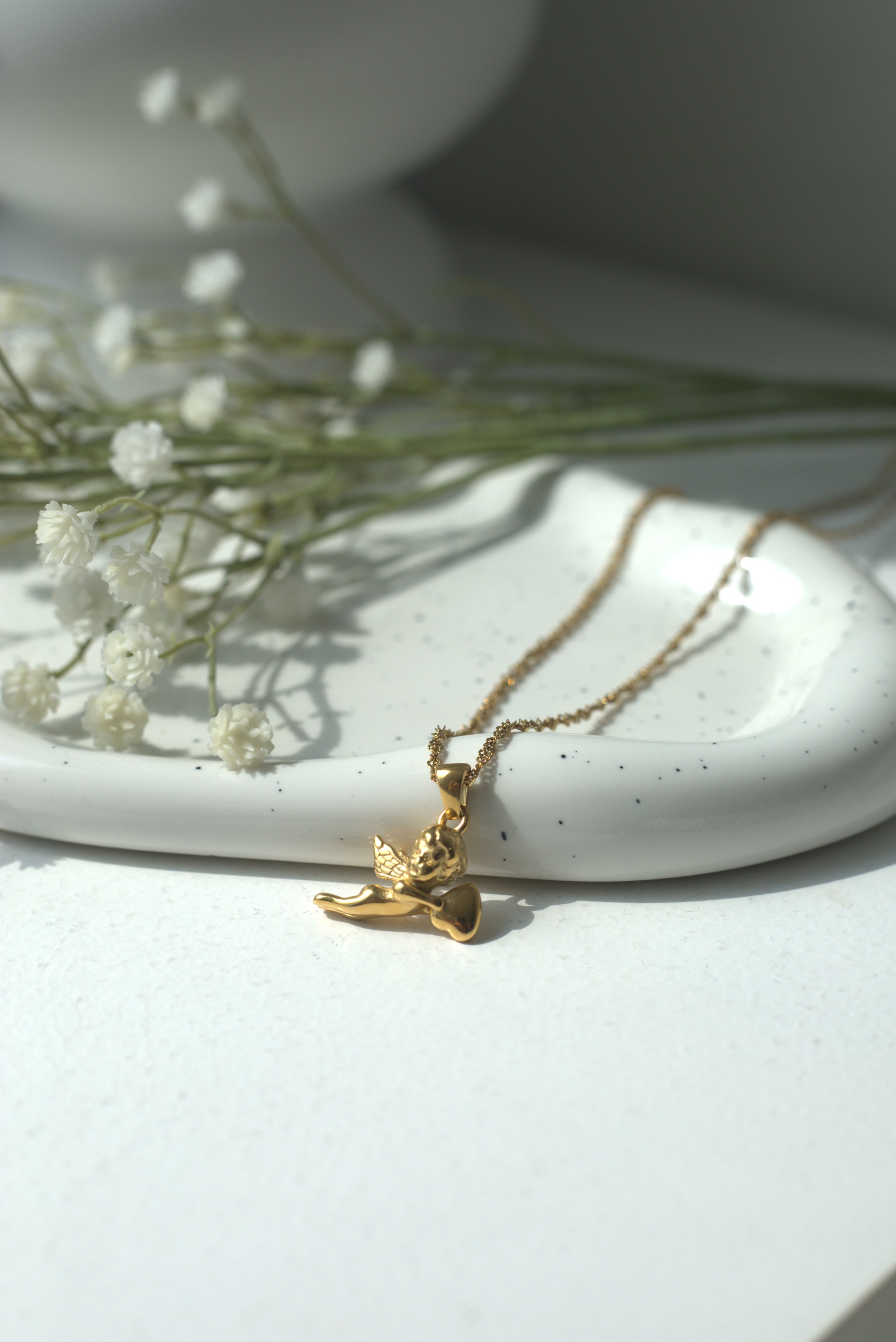 Gold necklace with a small cupid pendant on a white surface with dried flowers.