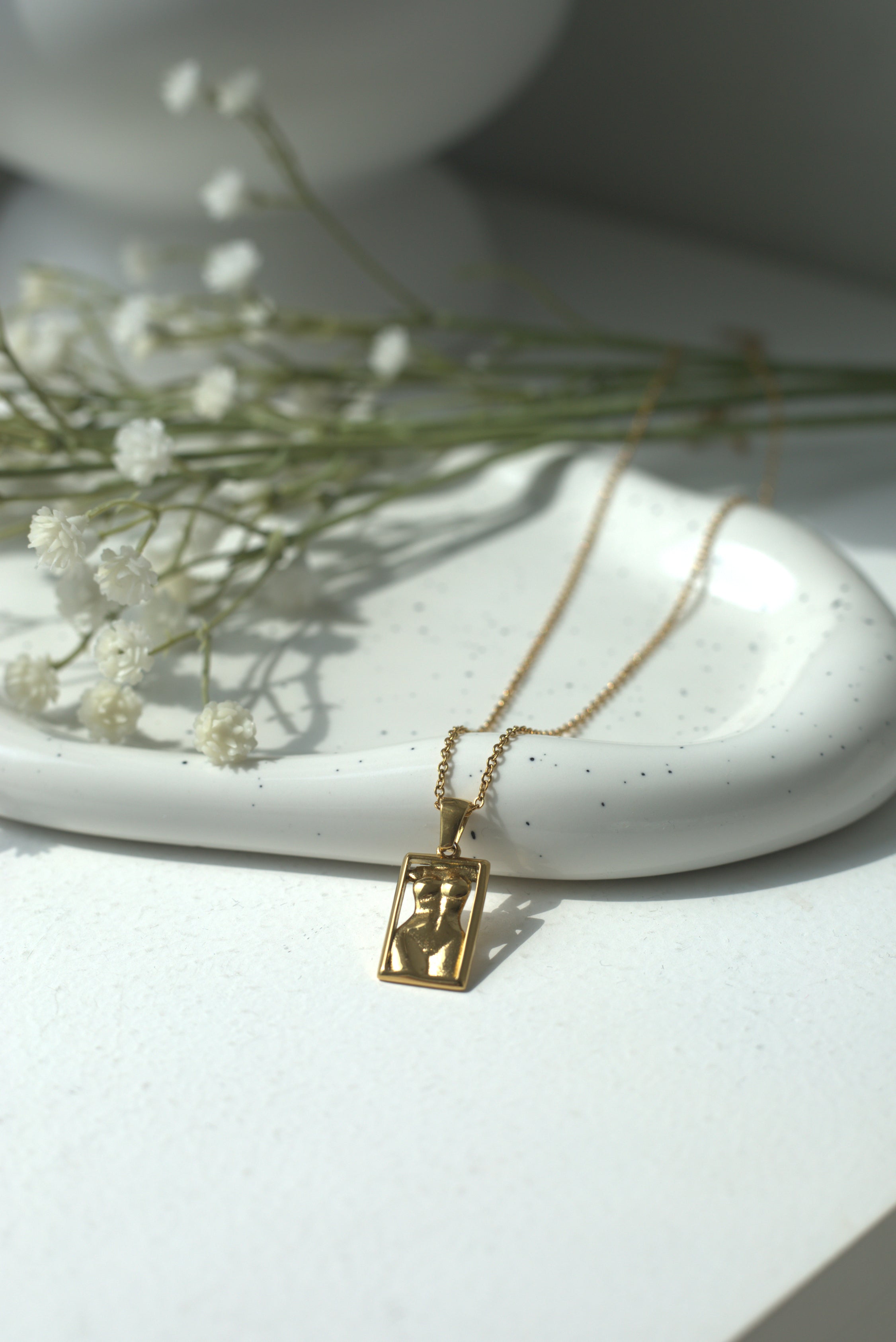 Gold necklace with a rectangular women's silhouette pendant on a white surface with dried flowers.