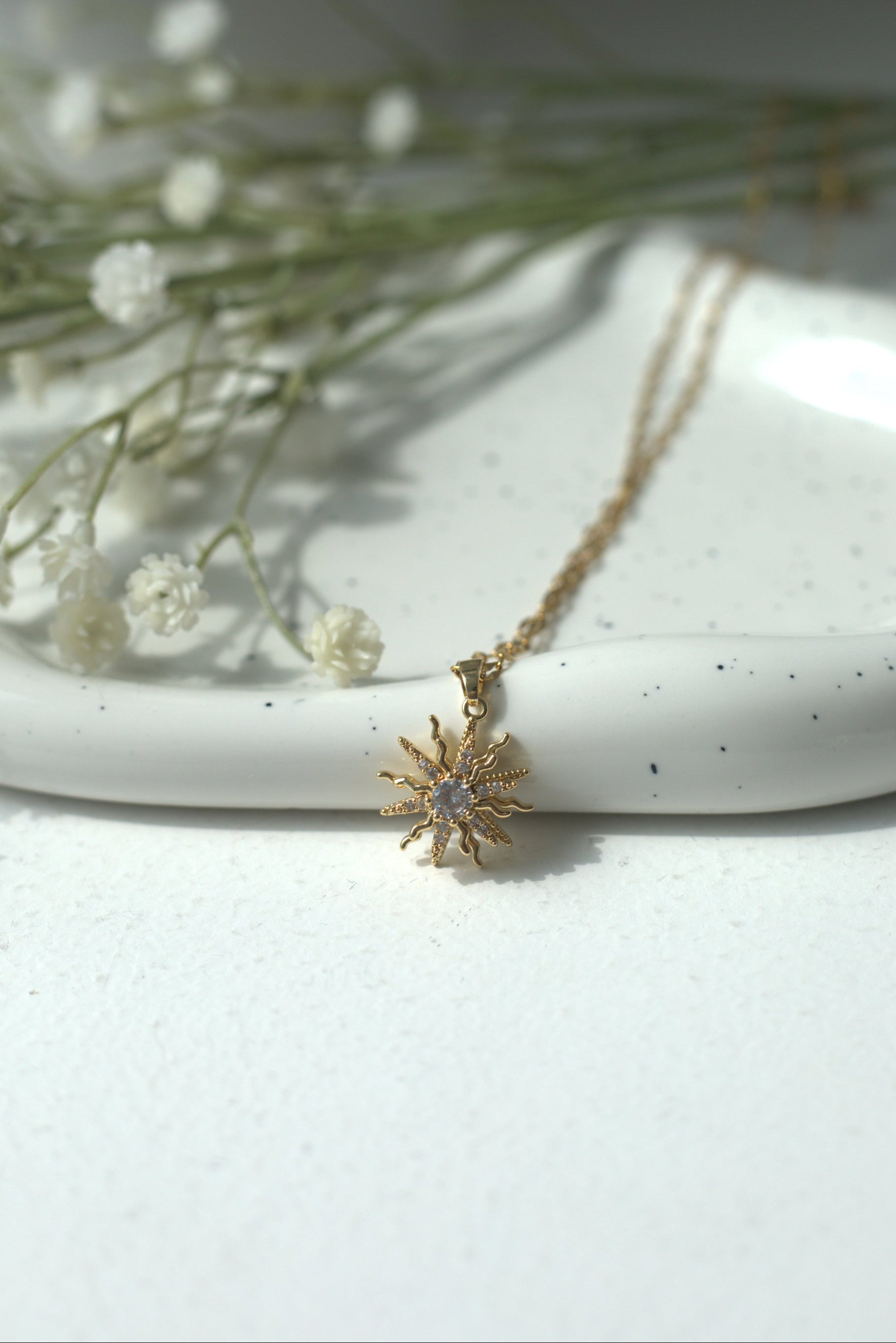 Gold waterproof sunny necklace with a star-like pendant on a white surface with dried flowers.