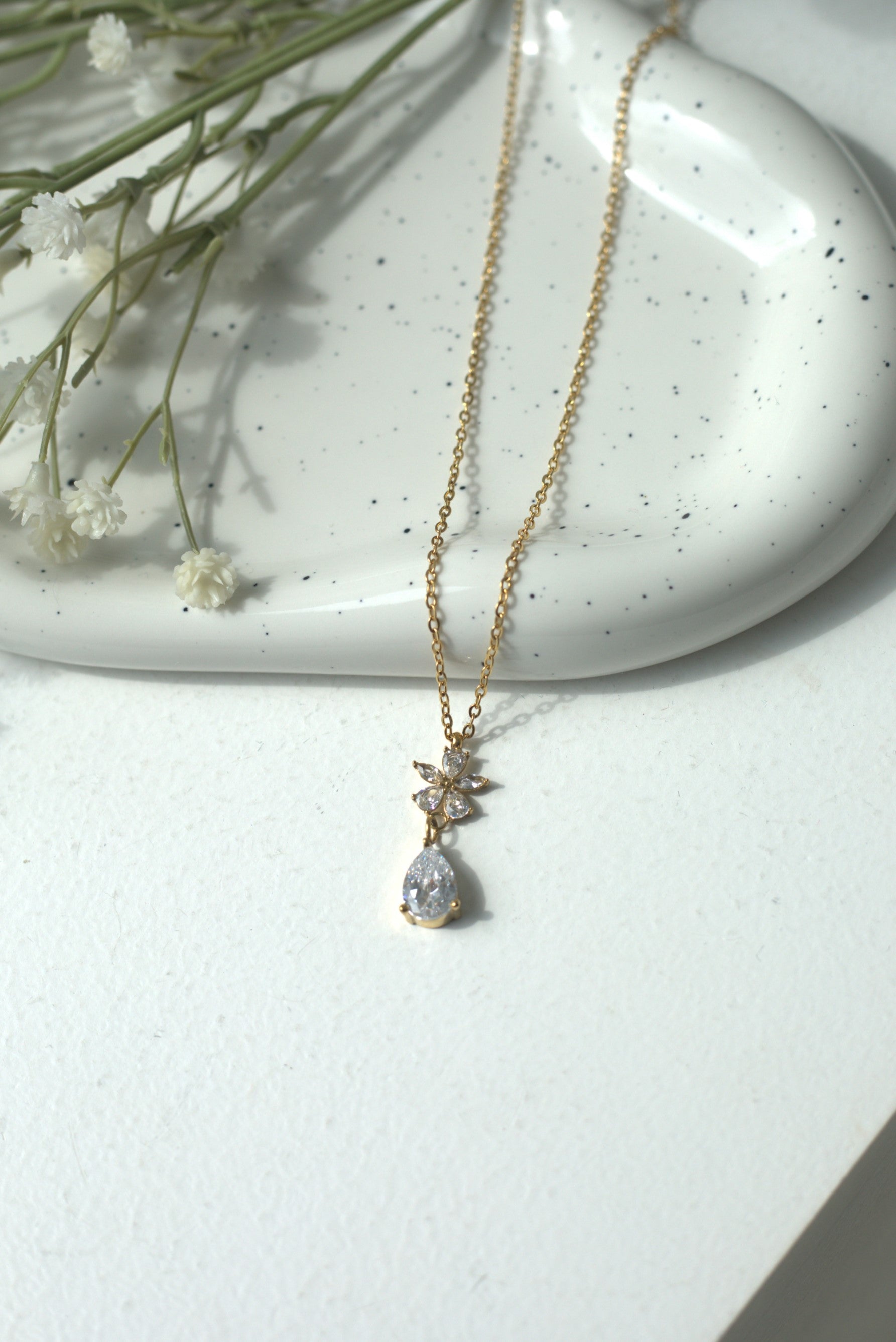 Delicate gold necklace with a teardrop pendant on a white surface with small flowers.