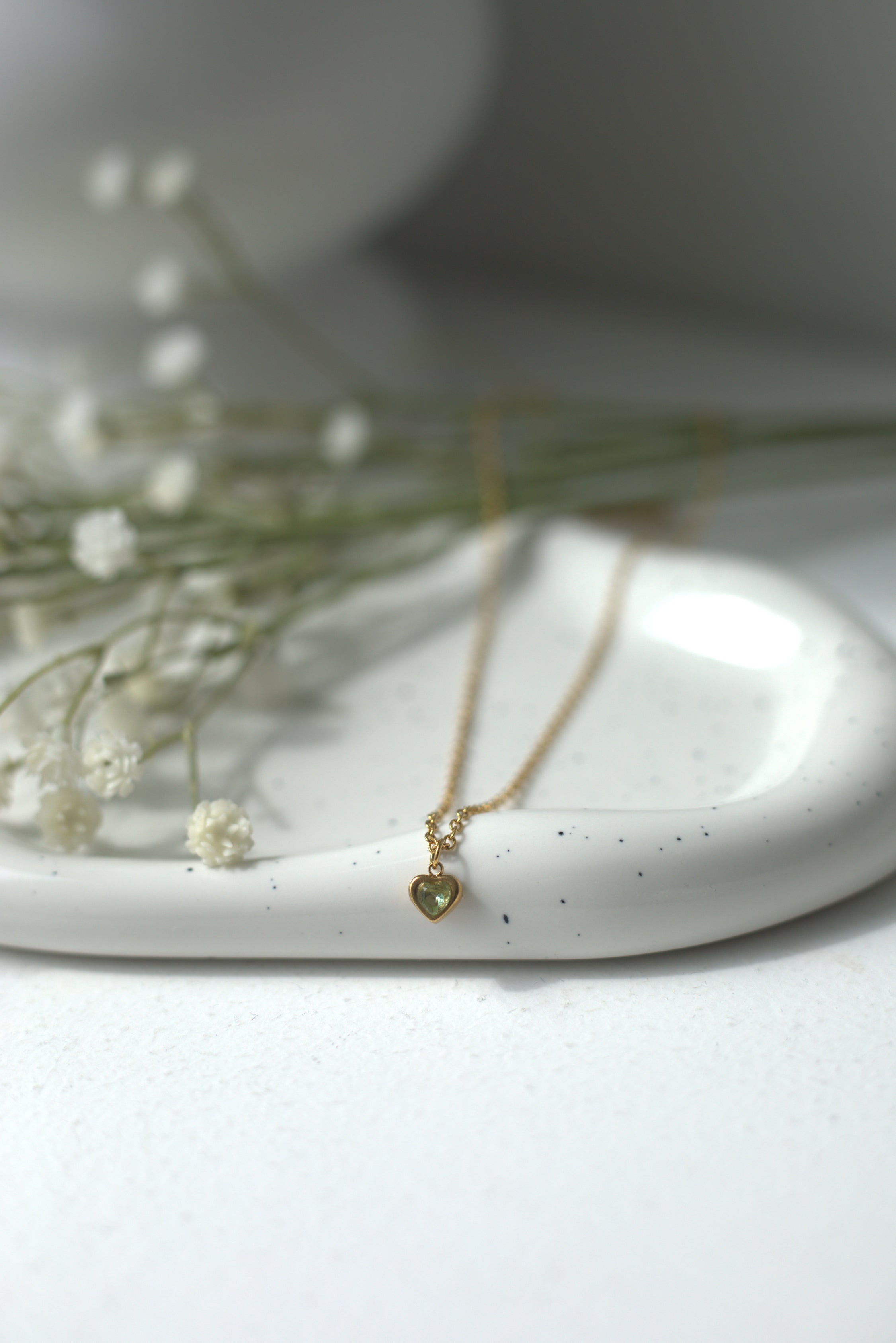 Delicate gold necklace with a heart-shaped pendant on a white ceramic dish with small flowers.