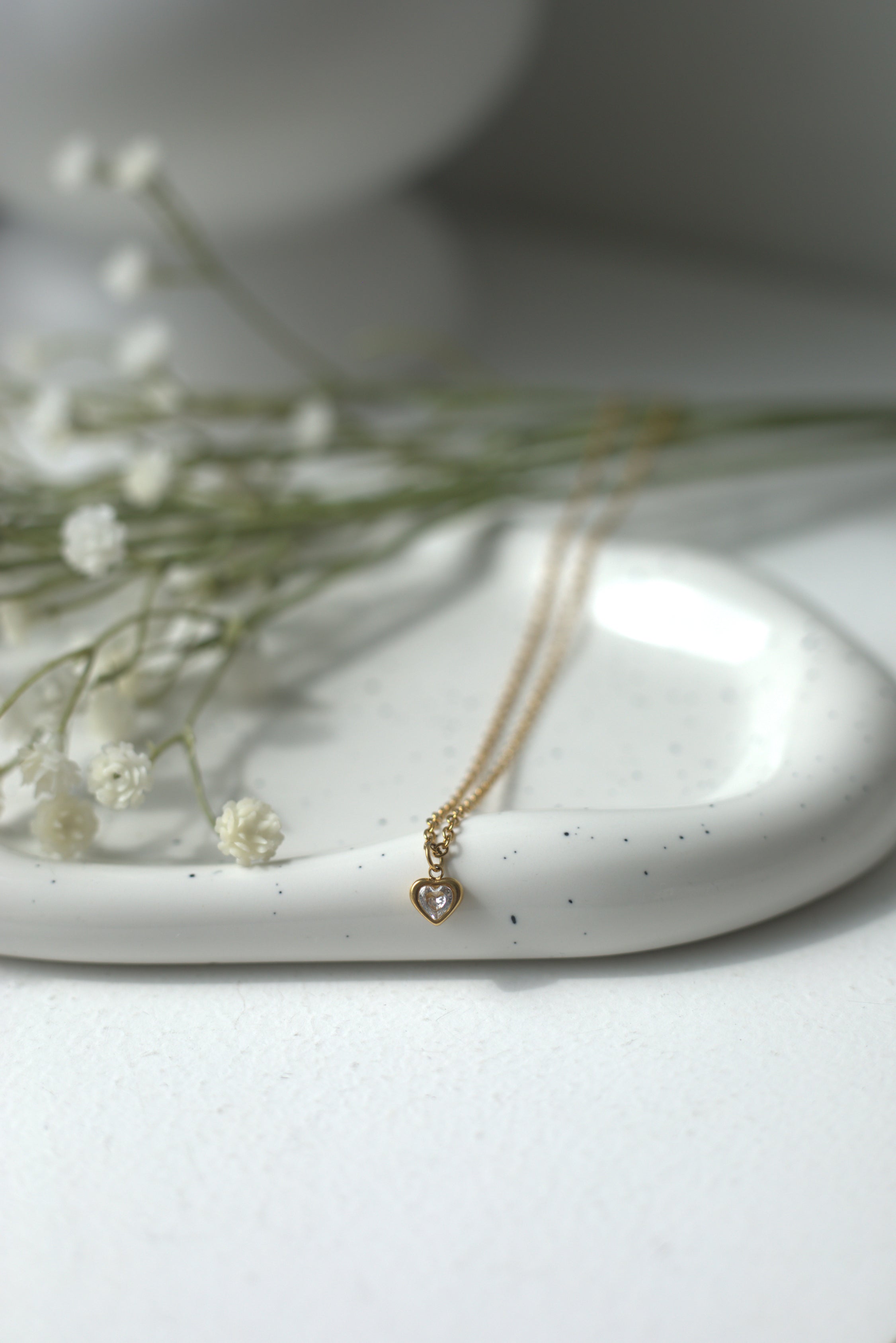 Gold necklace with a small white heart pendant on a white ceramic dish with dried flowers.