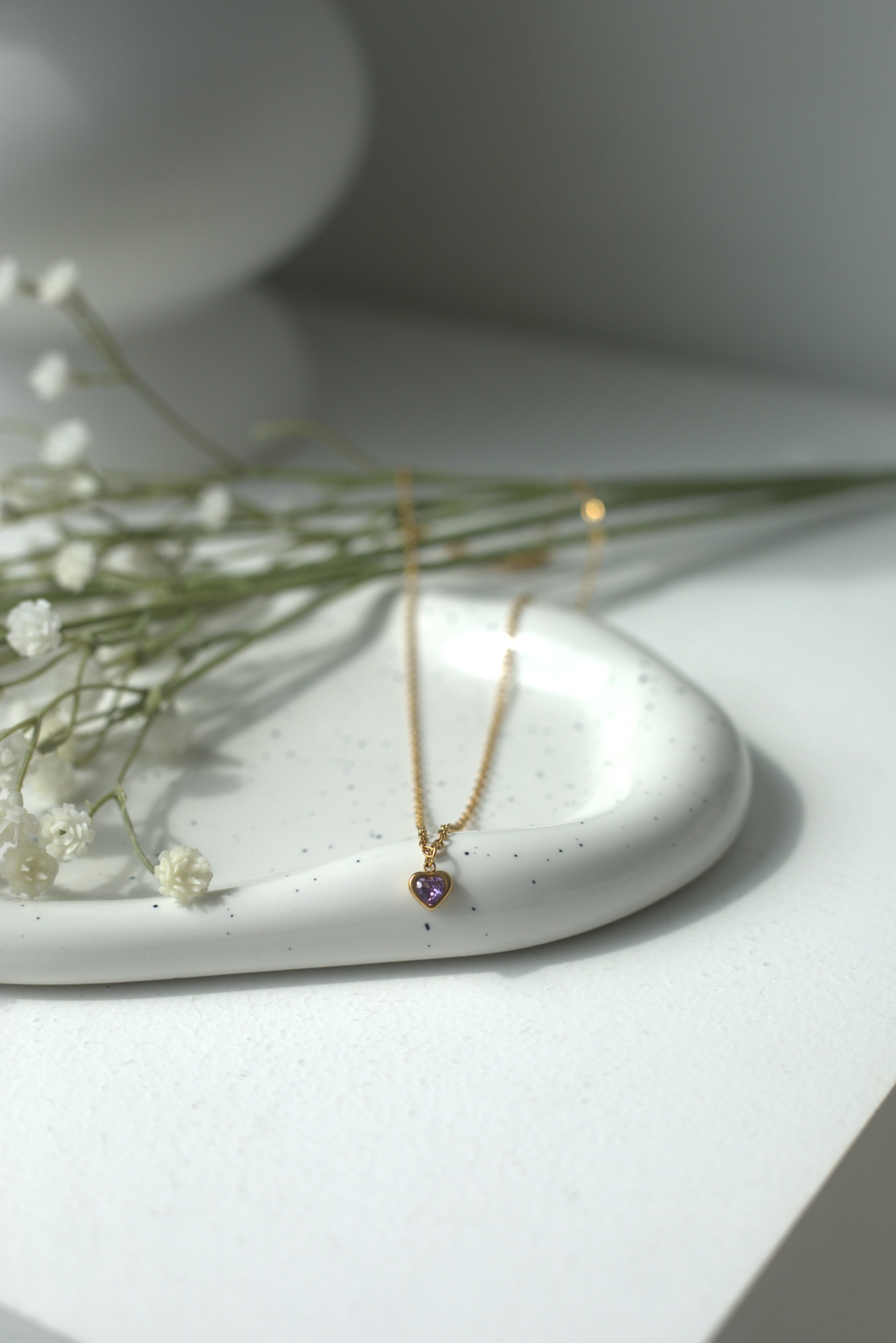 Gold necklace with a purple heart pendant on a white speckled dish with greenery