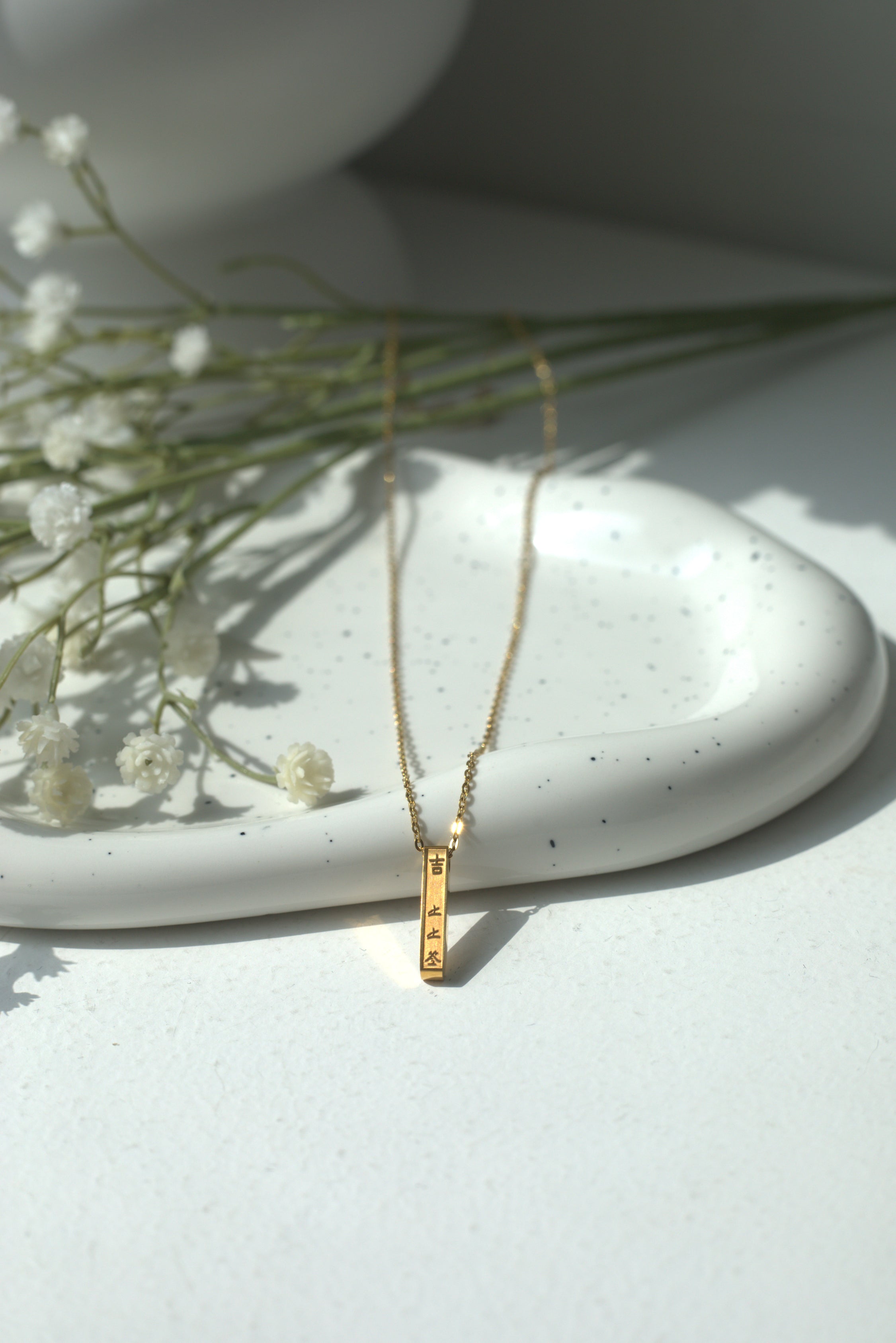 Gold bar necklace with a Chinese lucky message on a white surface with small white flowers