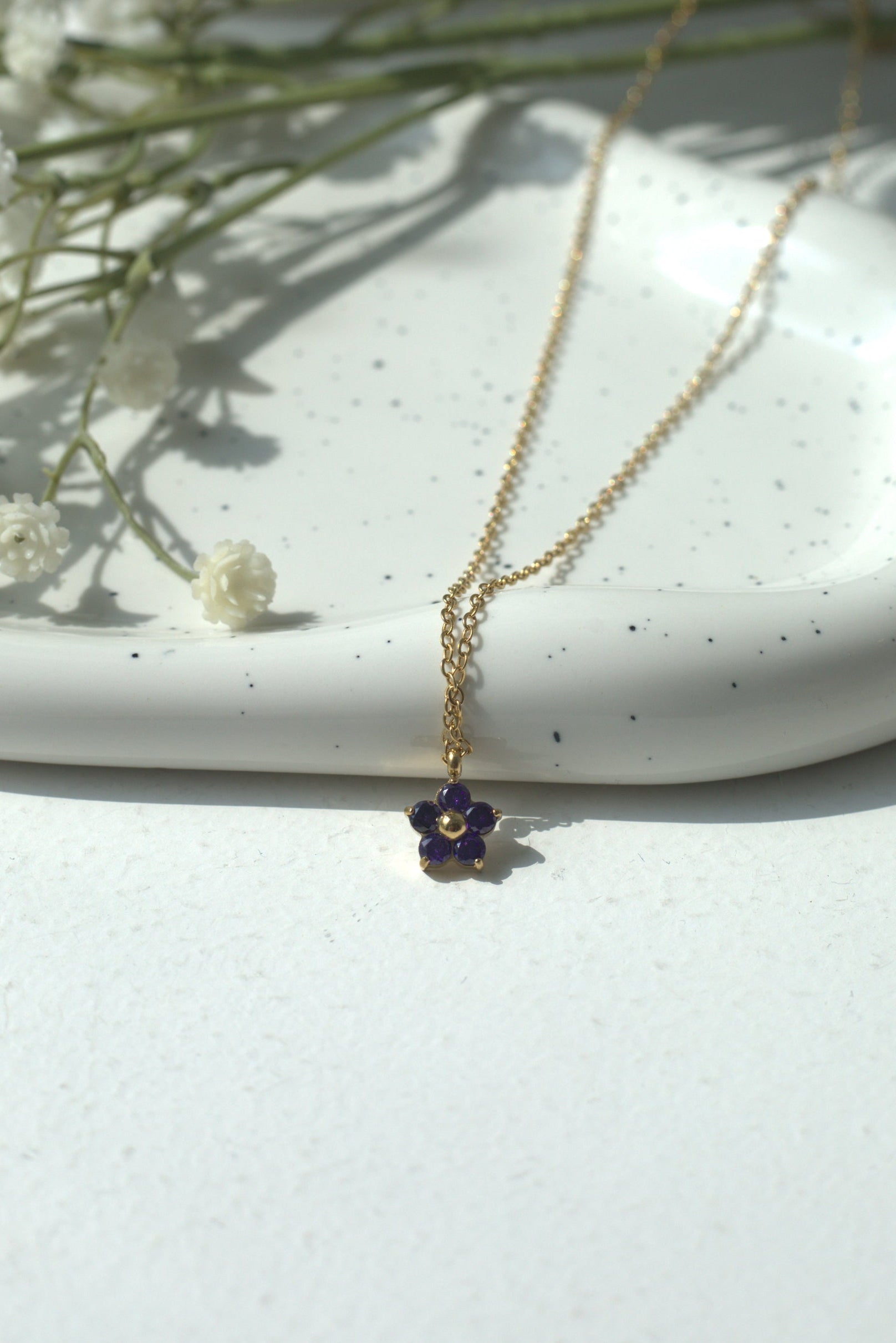 Gold waterproof necklace with a small purple pendant on a white surface with baby's breath flowers.
