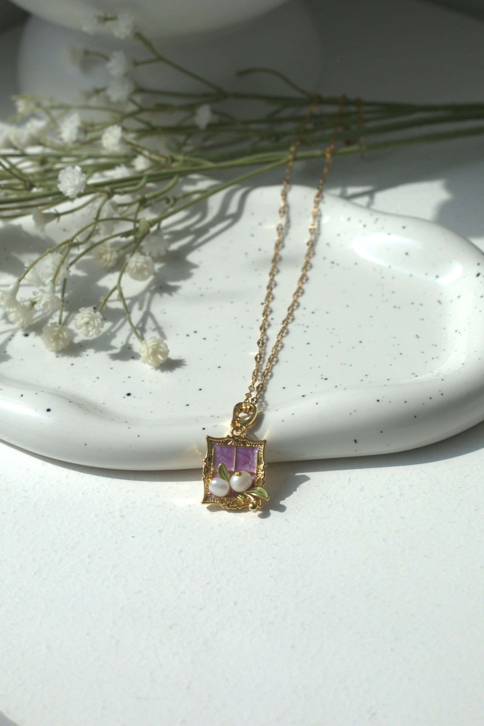 Gold necklace with a purple gemstone pendant on a white surface with flowers.