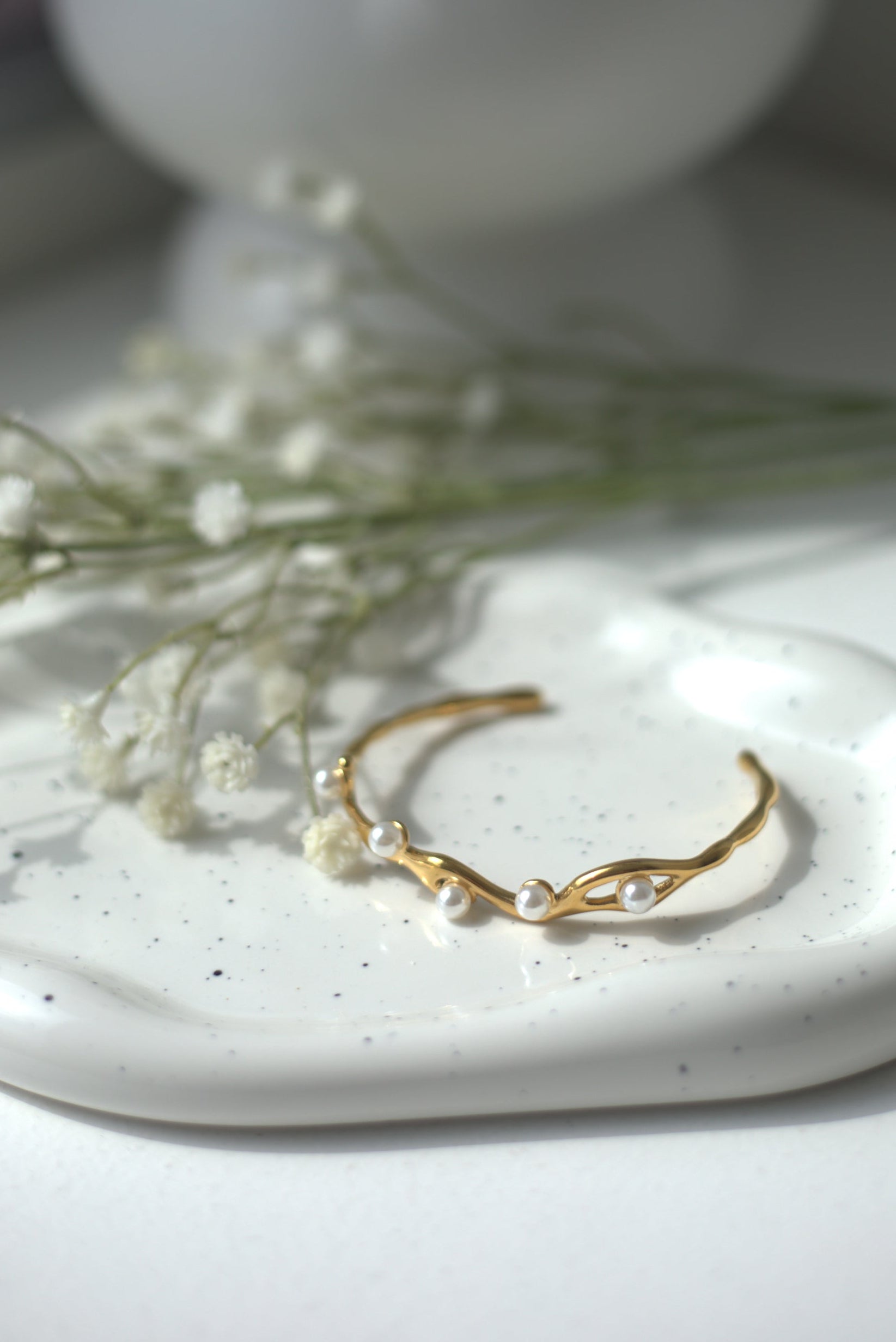 Gold bracelet with pearl accent on a white ceramic dish with small flowers and a white vase in the background