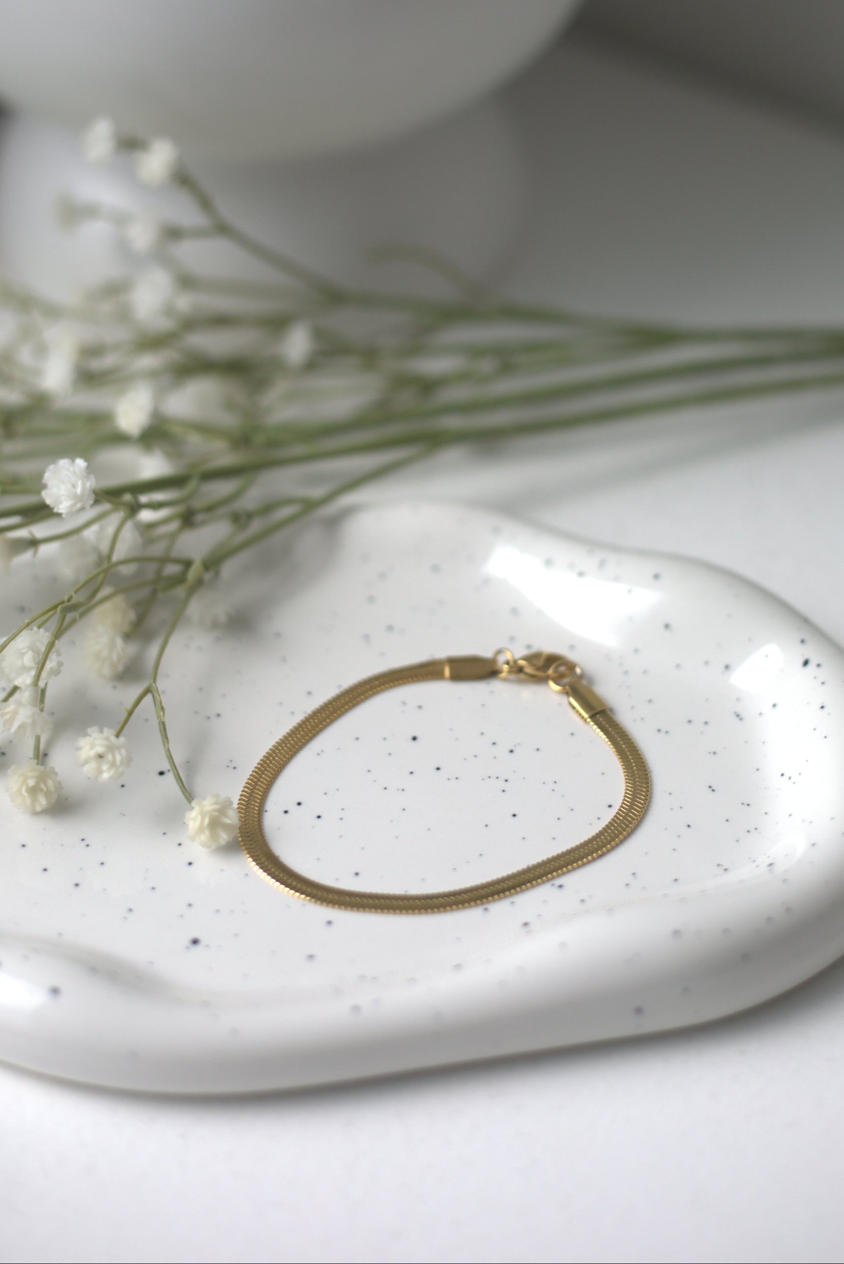 Gold bracelet on a white speckled dish with small white flowers and a white vase in the background.