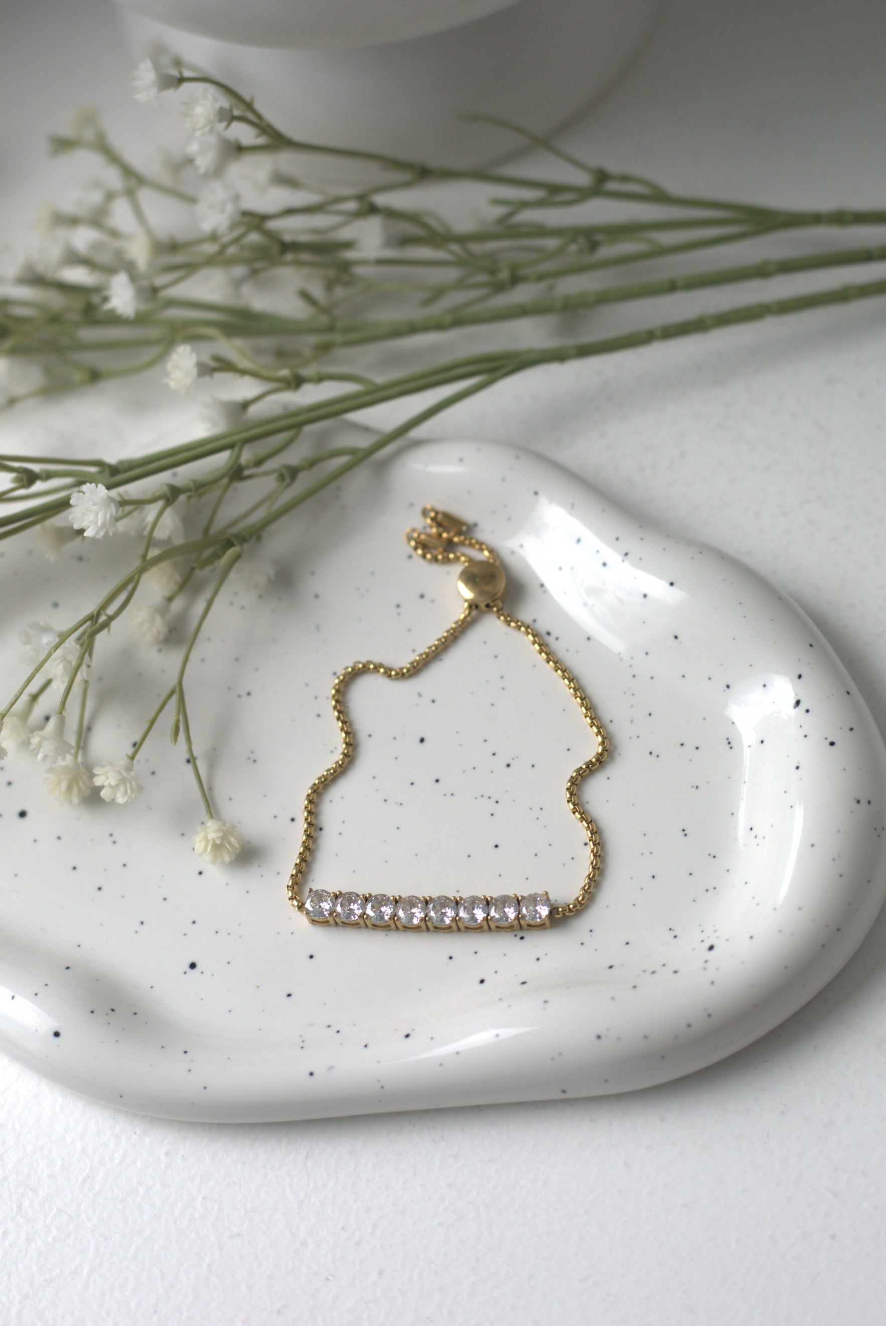 Gold bracelet on a white speckled dish with small flowers and a white vase in the background.
