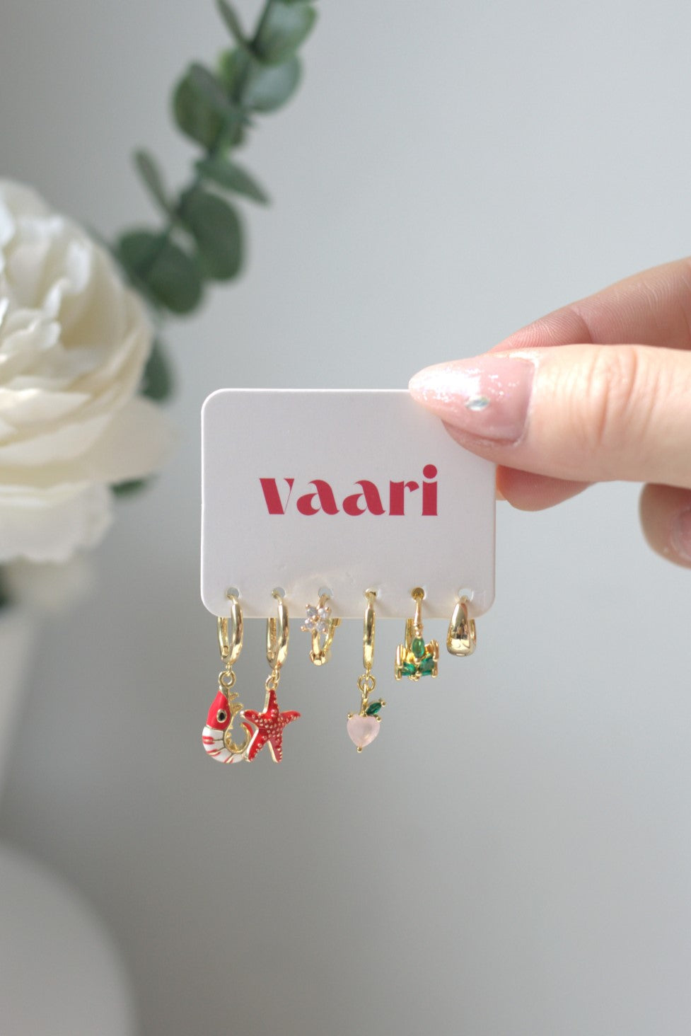 Set of gold earrings on a 'Vaari' branded card held by a hand with a white flower in the background.