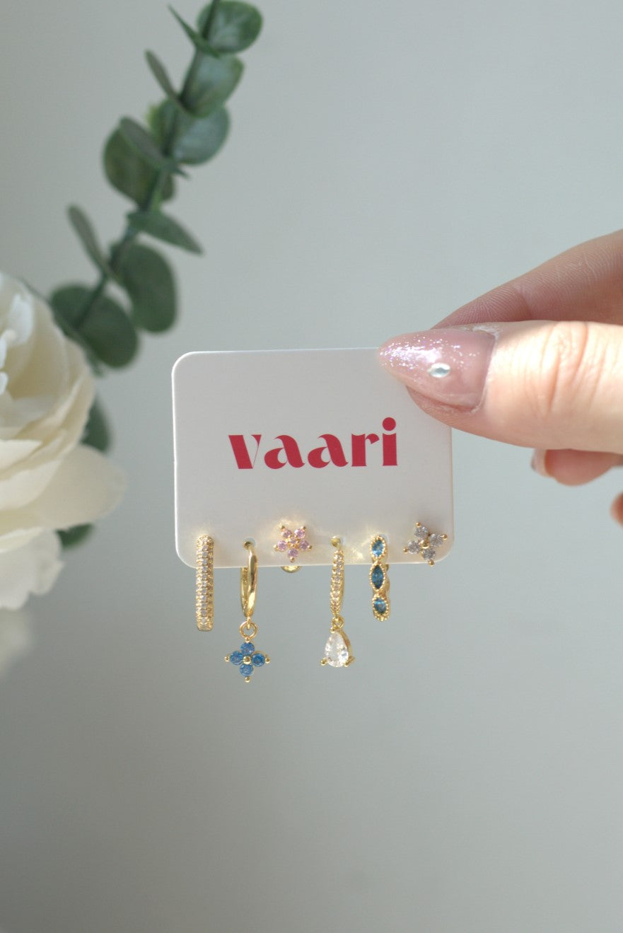 Hand holding a card with gold earrings with light blue and white gemstones on a white background, featuring the brand 'Vaari'.