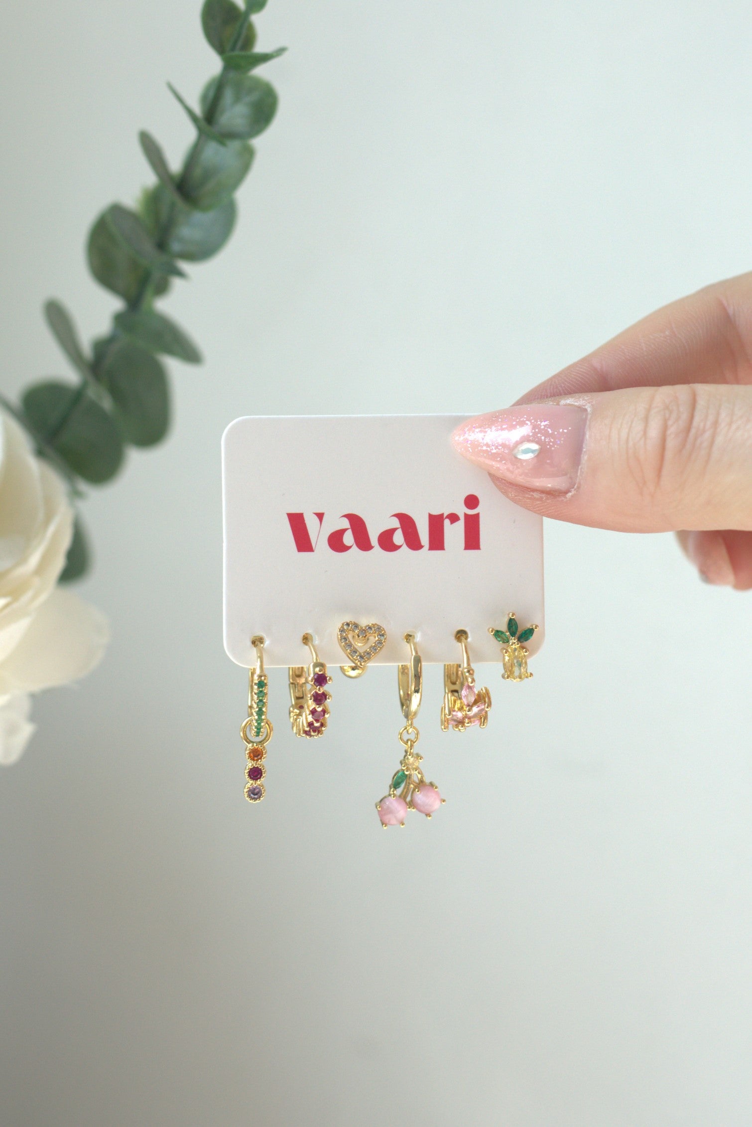 Hand holding a card with earrings in heart, pineapple, cherry designs on a white background