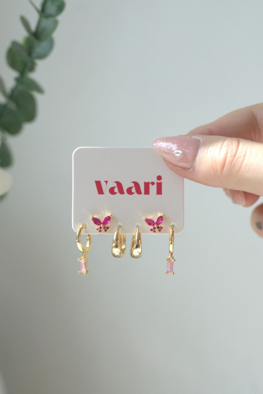 Gold and pink stainless steel butterfly earrings on a 'vaari' branded card held by a hand.