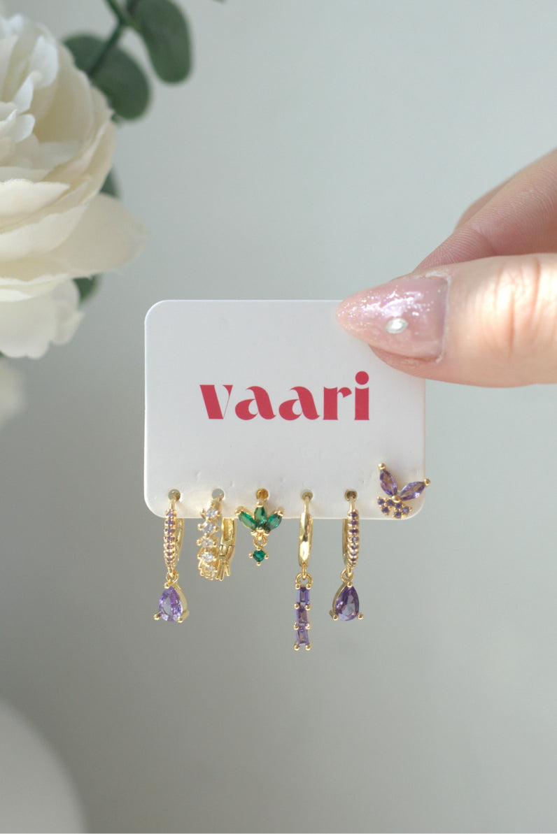 Set of gold and purple / green dangly earrings on a 'vaari' branded card with a white flower in the background