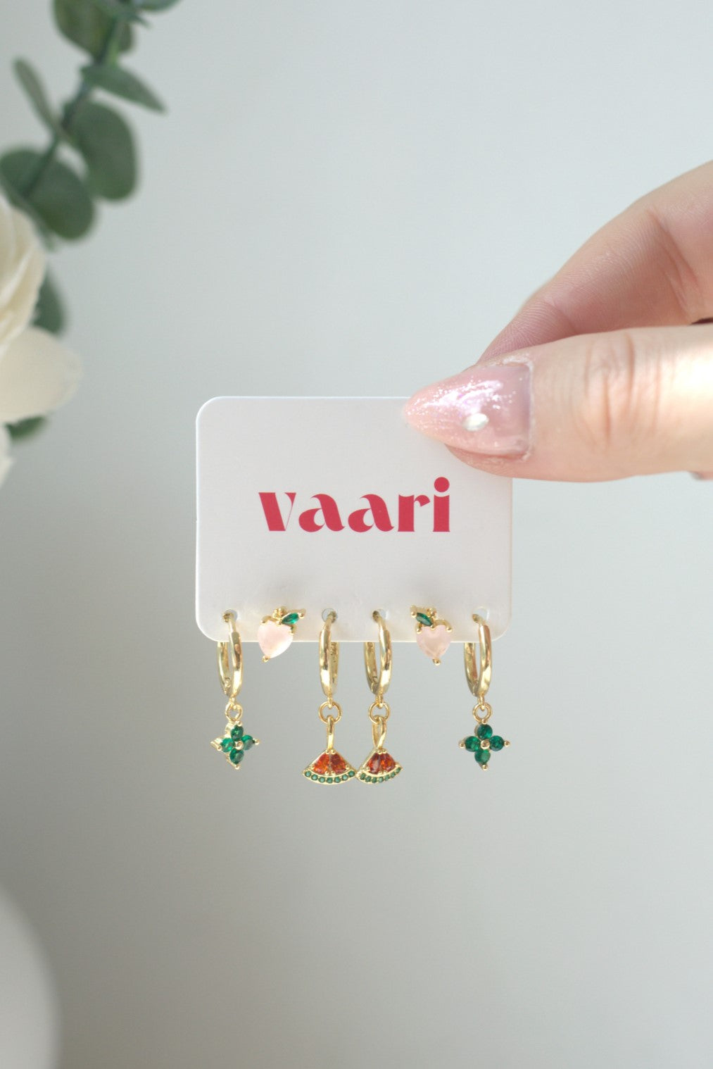 Gold stainless steel earrings with fruit themed gemstones on a 'vaari' branded card held by a hand.