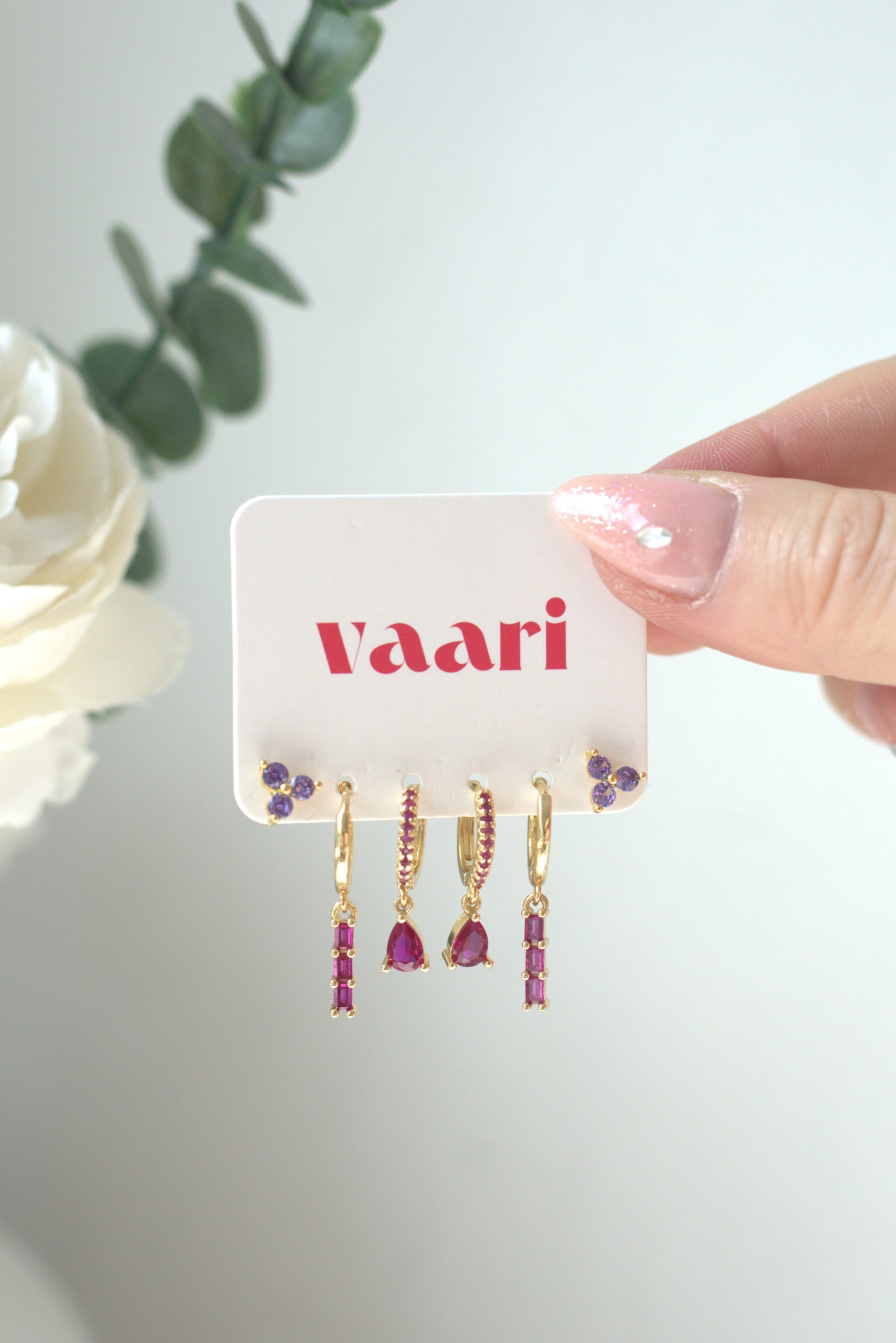 Hand holding a card with 'vaari' branding and gold & dark pink dangle earrings on a light background