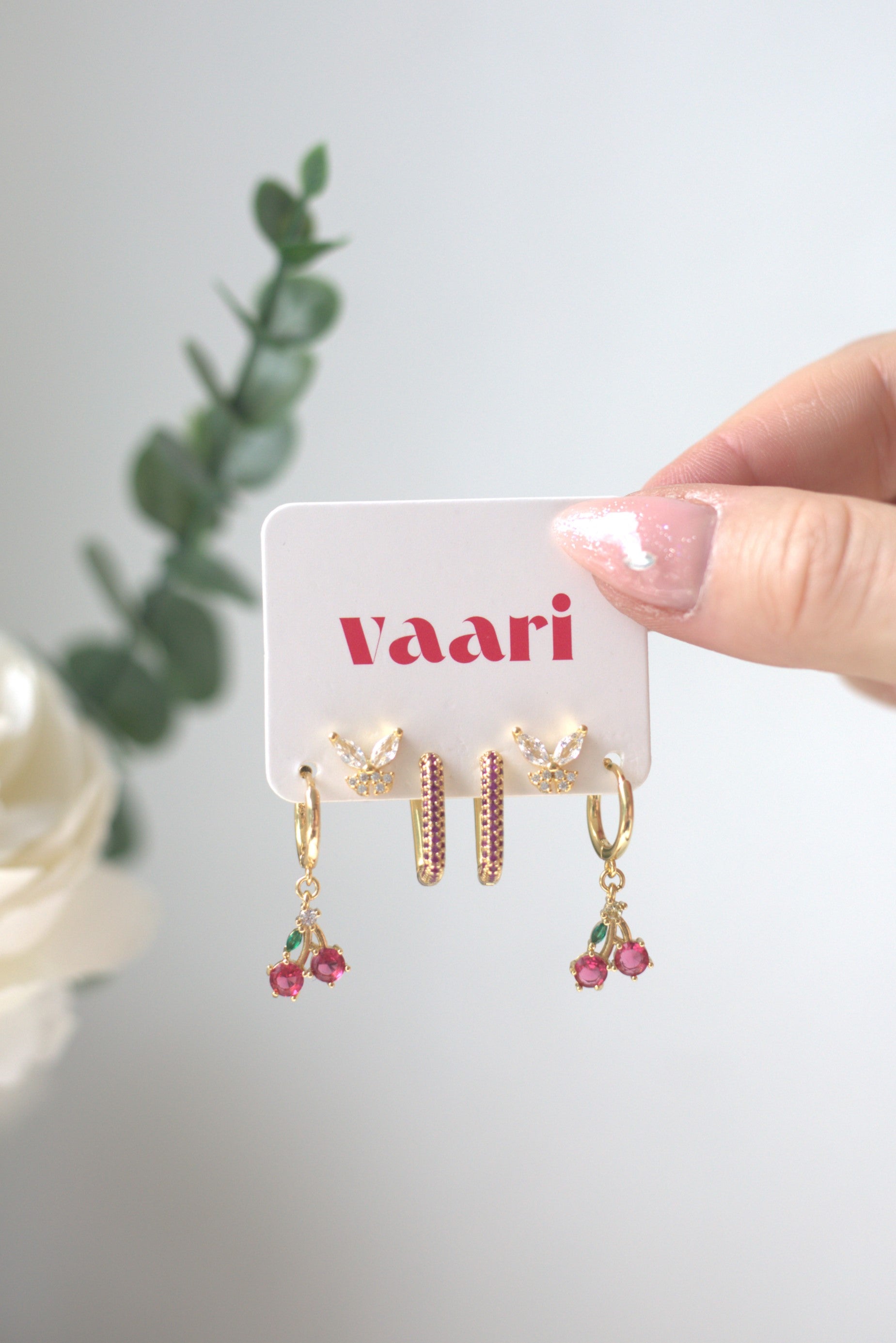 Gold square and cherry hoop earrings with red pinkish accents on a 'Vaari' branded card, held by a hand against a light background. Butterfly studs