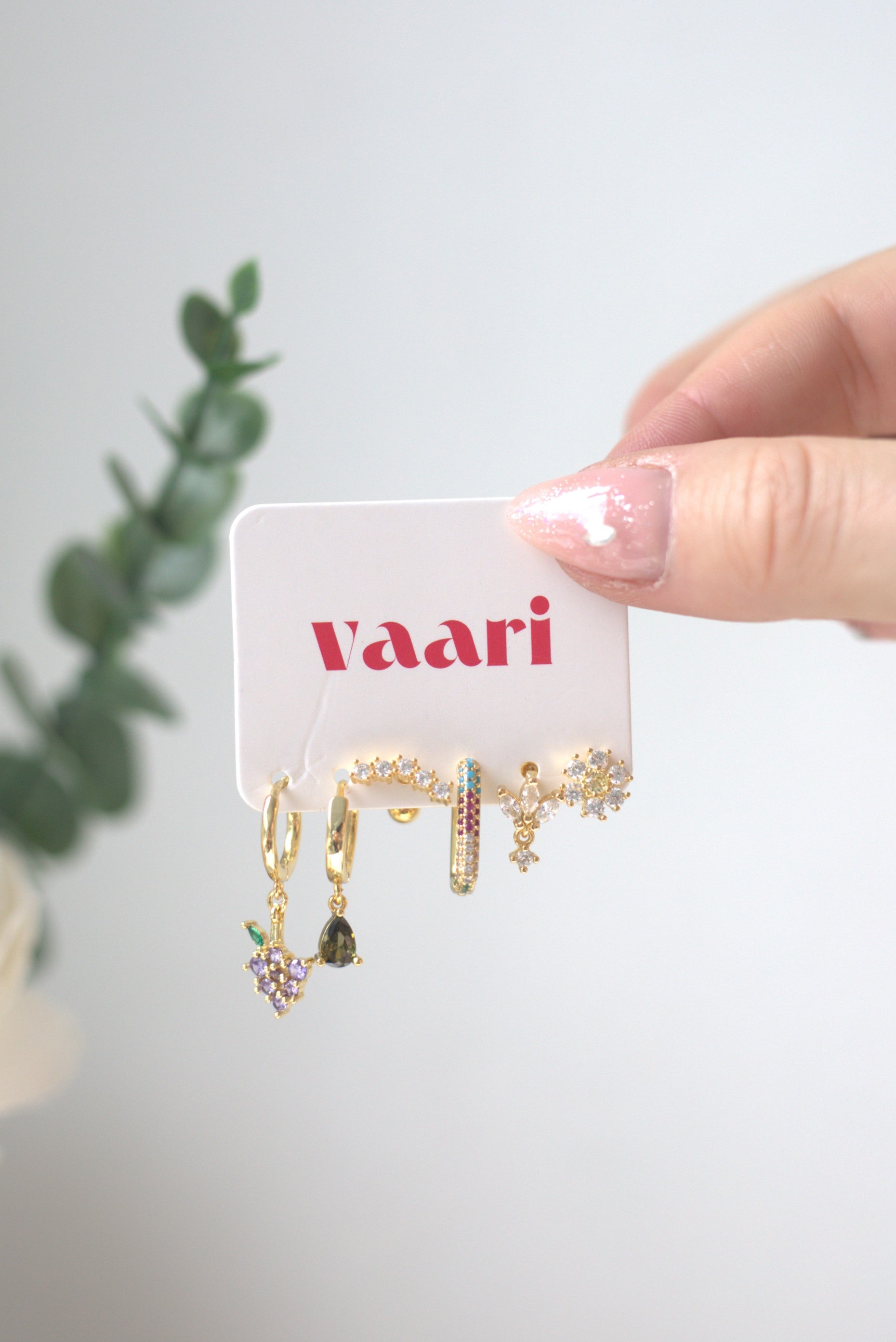Gold earrings with colorful stones on a 'vaari' branded card, held by a hand against a light background.