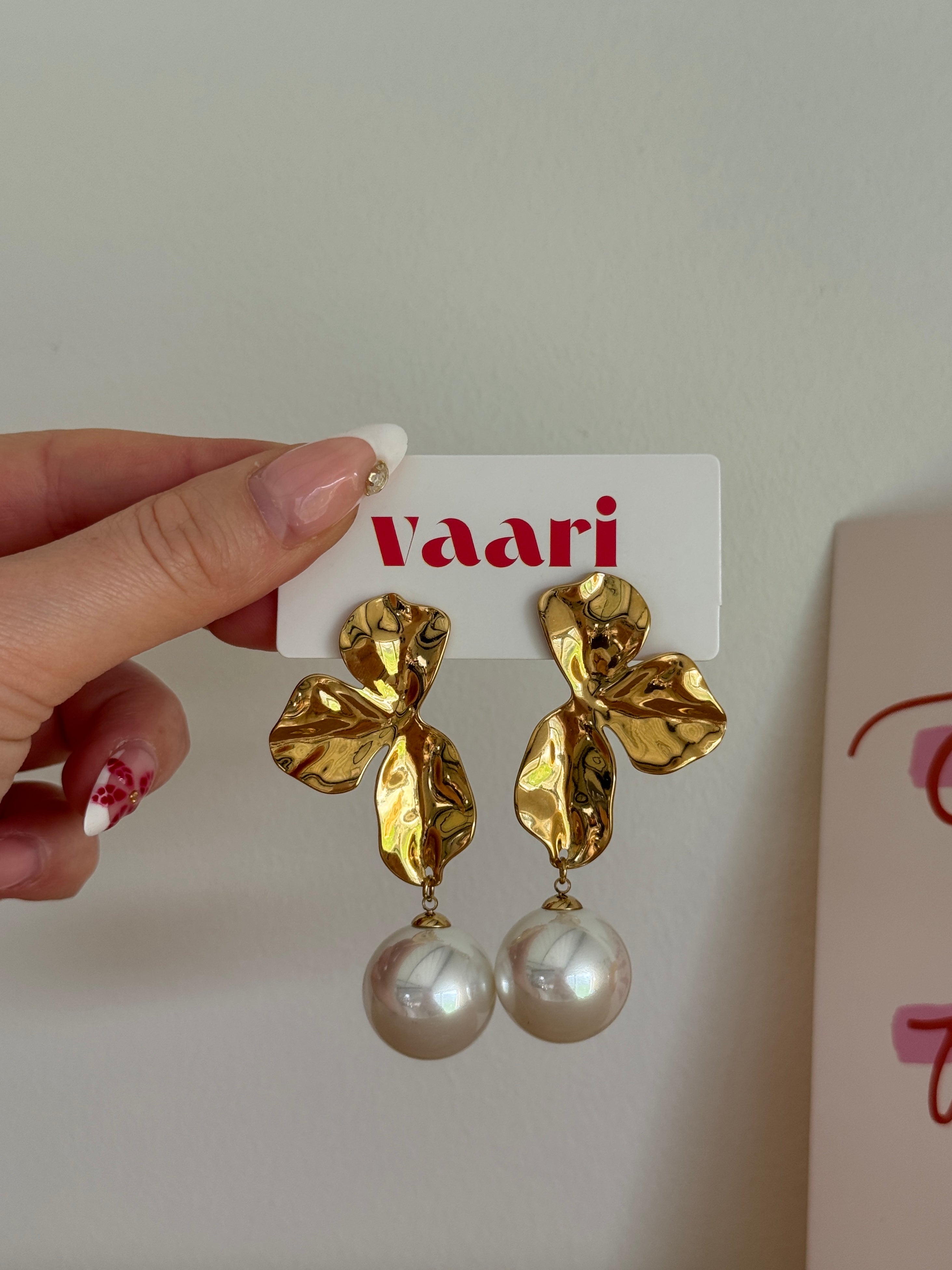 Gold floral stainless steel earrings with faux pearl drops held by a hand on a white background