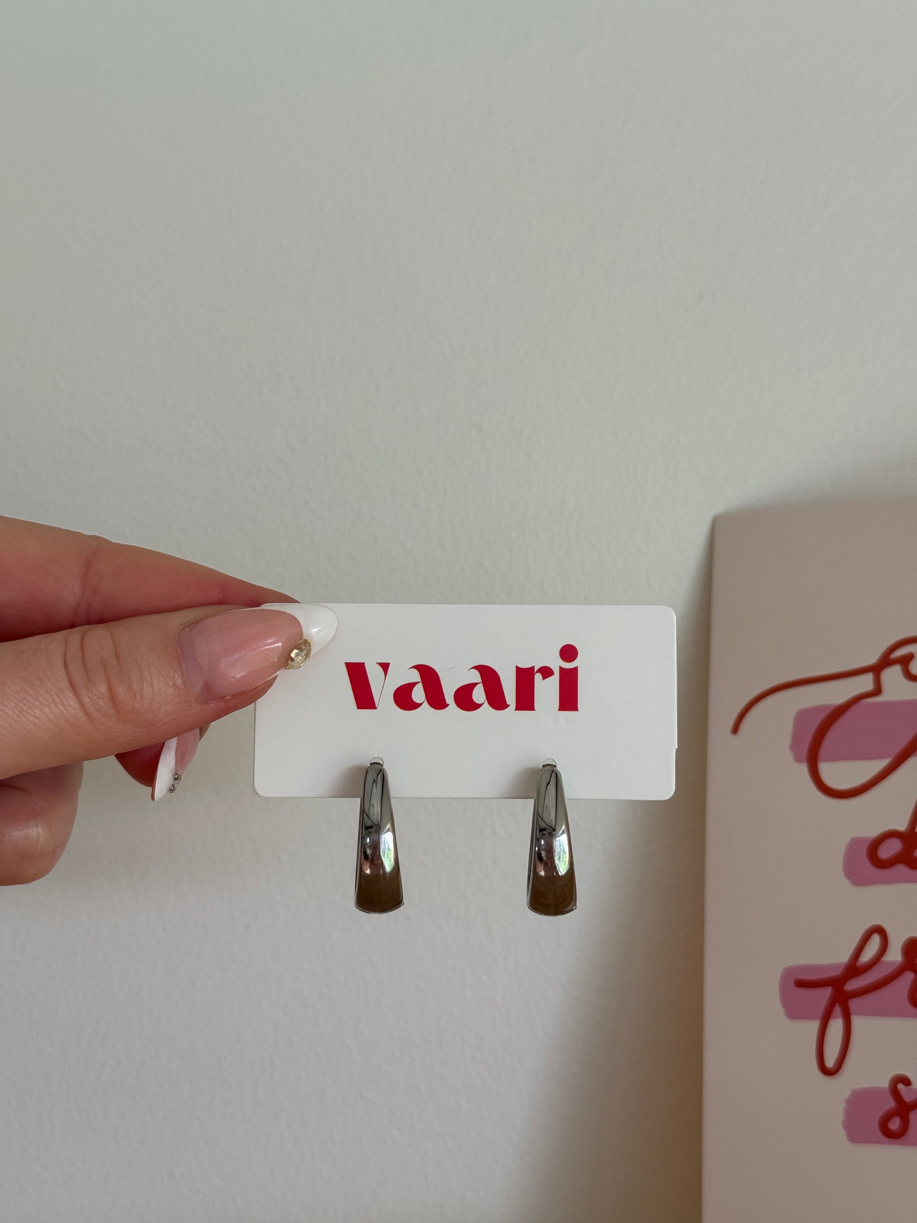Hand holding a card with 'Vaari' branding and stainless steel silver hoop earrings against a neutral background