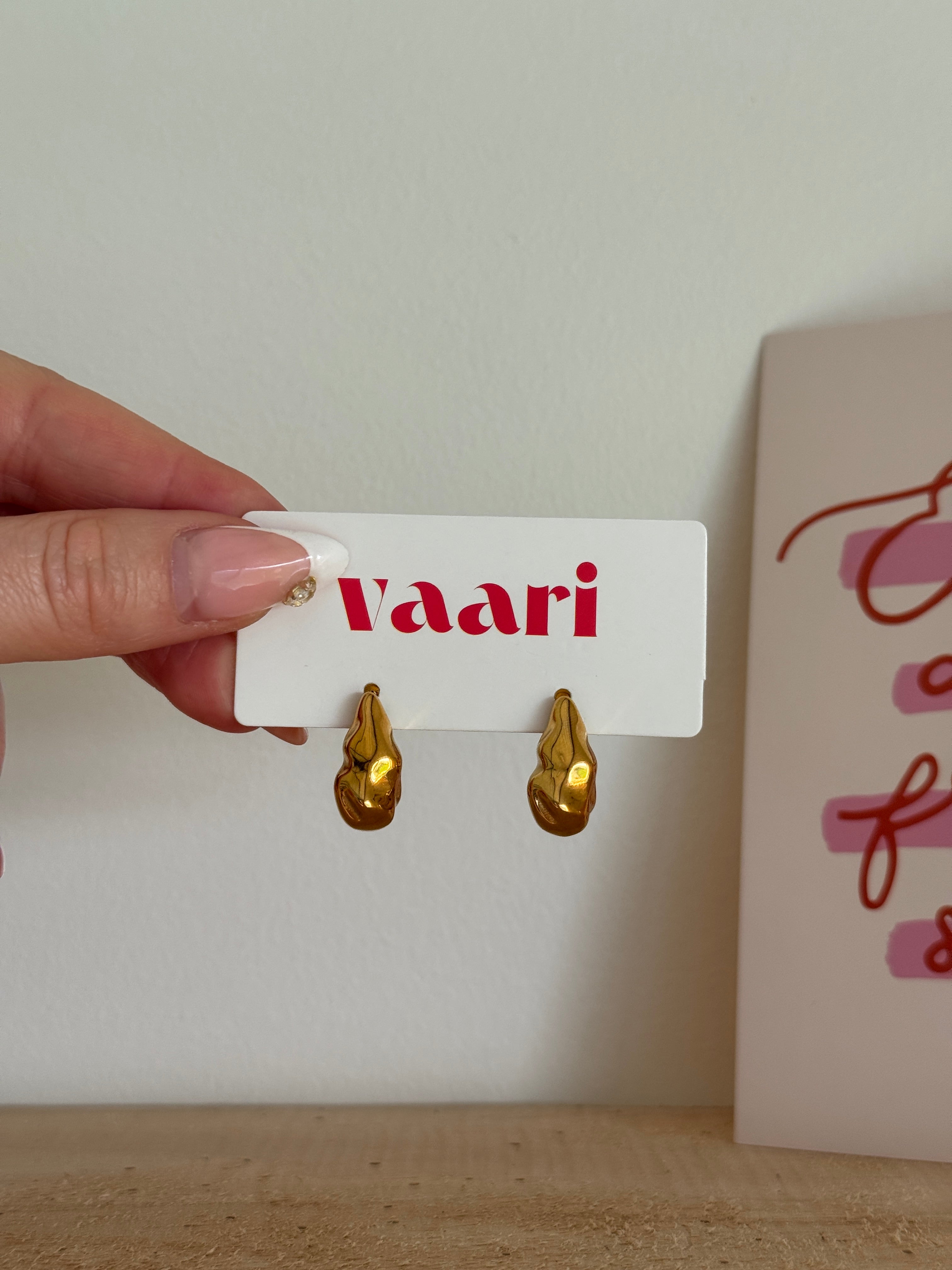 Gold earrings on a 'Vaari' card held by a hand against a neutral background