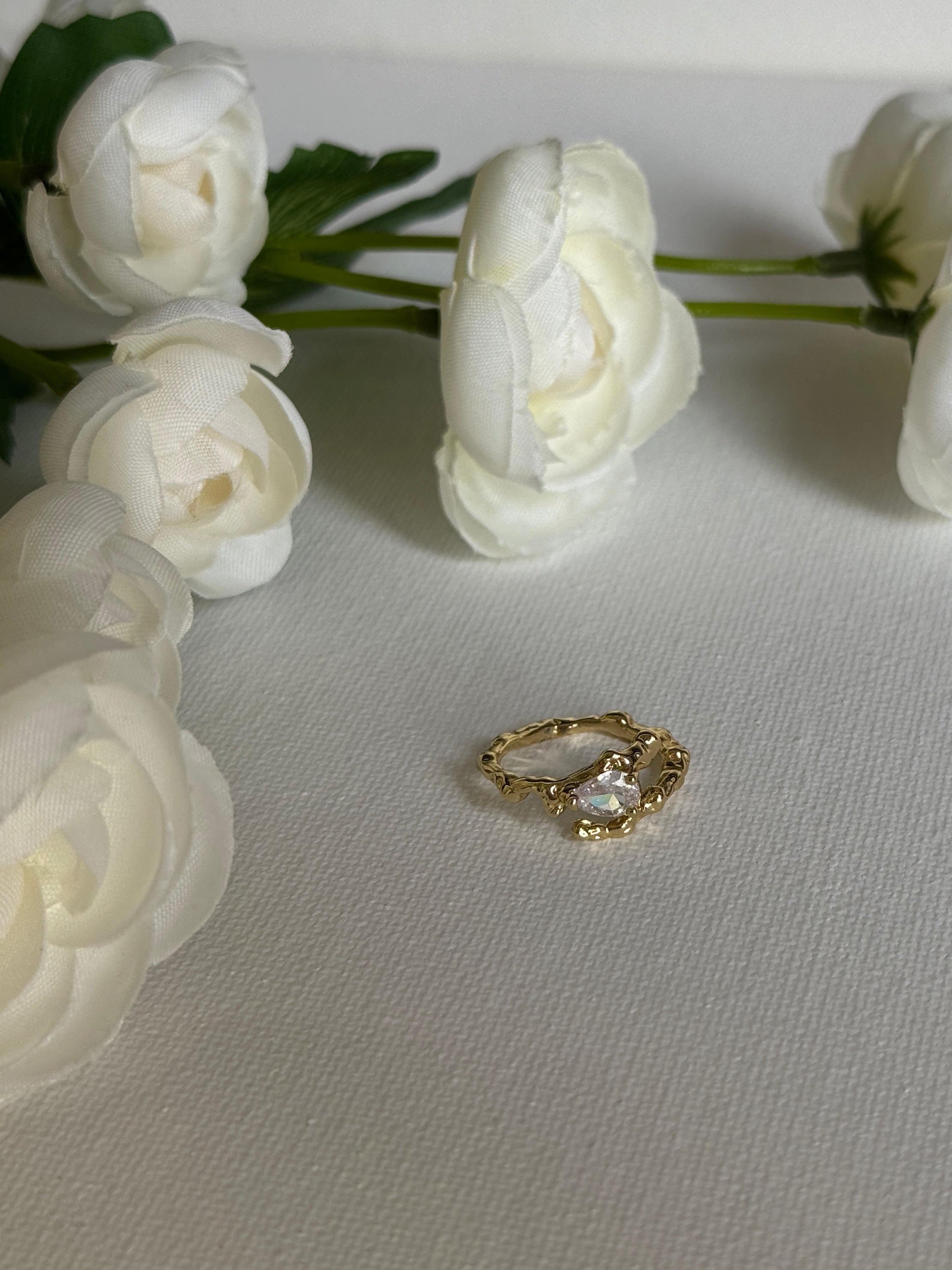 Gold hypoallergenic waterproof ring with a clear gemstone on a gray surface surrounded by white flowers.