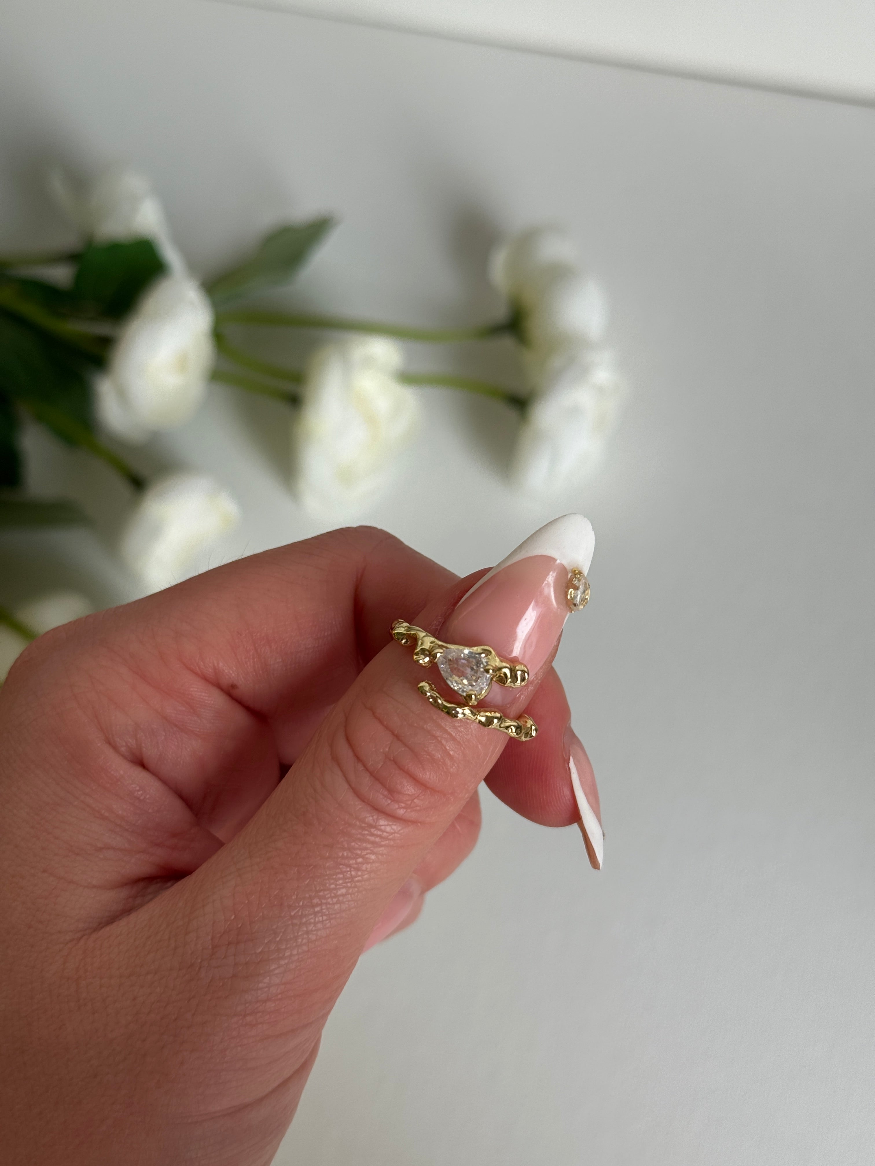 Gold size adjustable ring with a clear gemstone held by a hand against a white background with flowers.