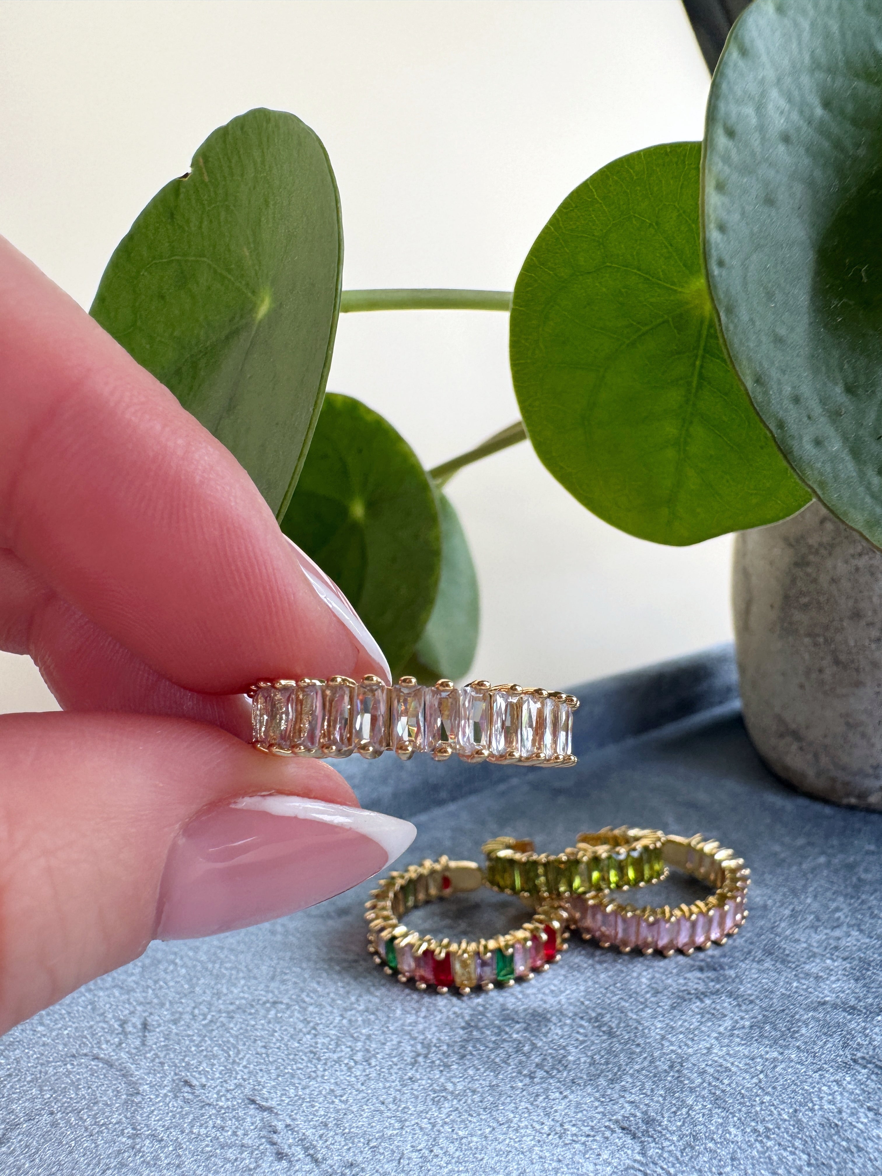 Hand holding a golden one size fits all ring with more rings on a textured surface and plant in the background