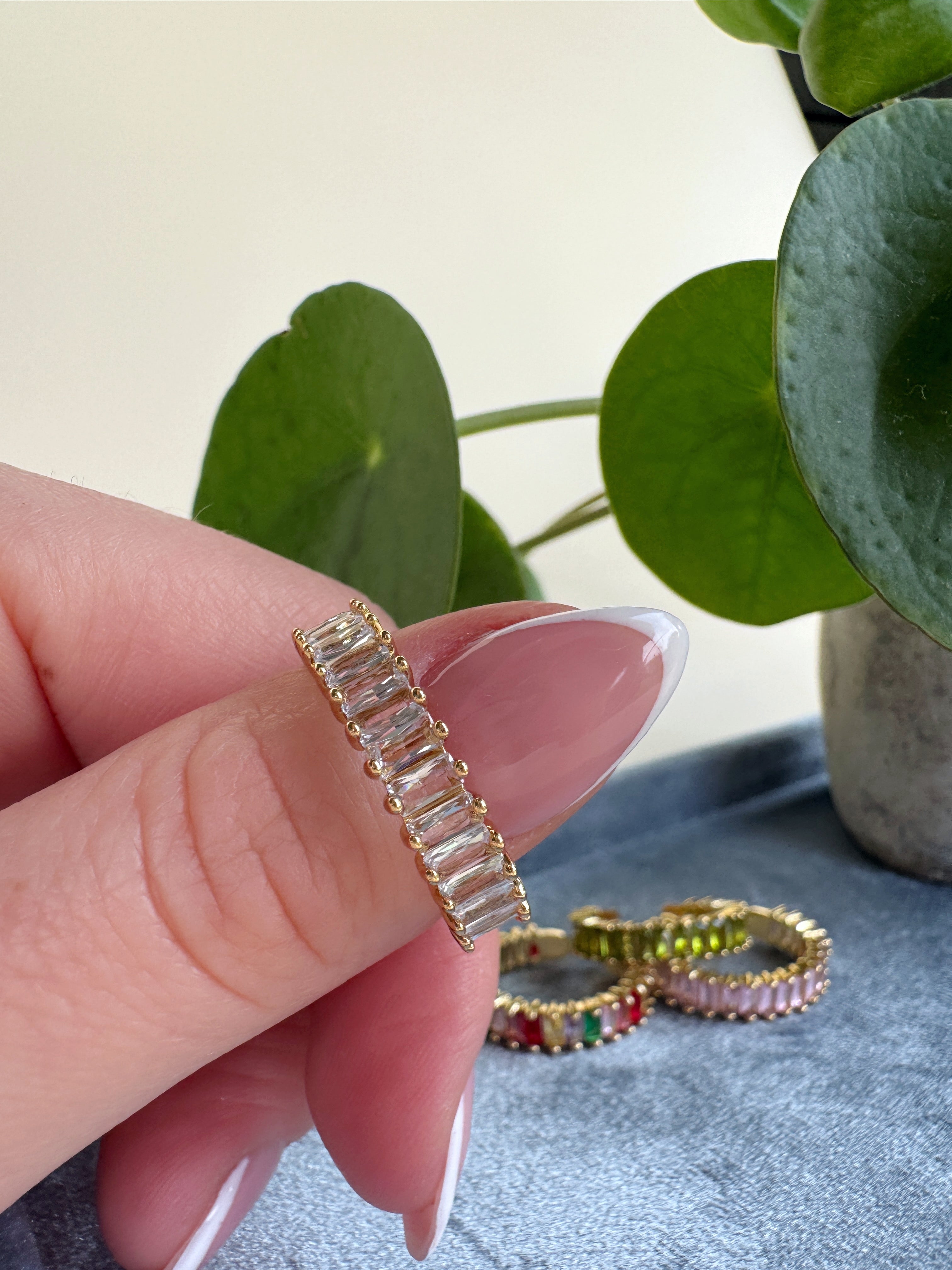 Gold ring with gemstones held by a hand, with more rings in the background on a textured surface.